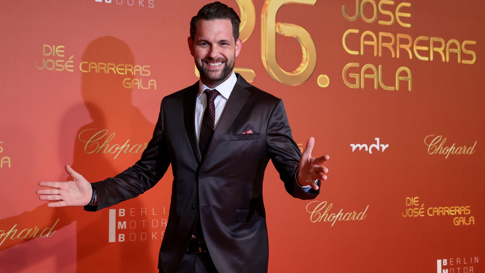 Moderator Matthias Killing kommt zur 26. Jose-Carreras-Gala. Das Bild zeigt ihn mit offenen Armen posierend vor einer Wand mit den Sponsoren der Gala.