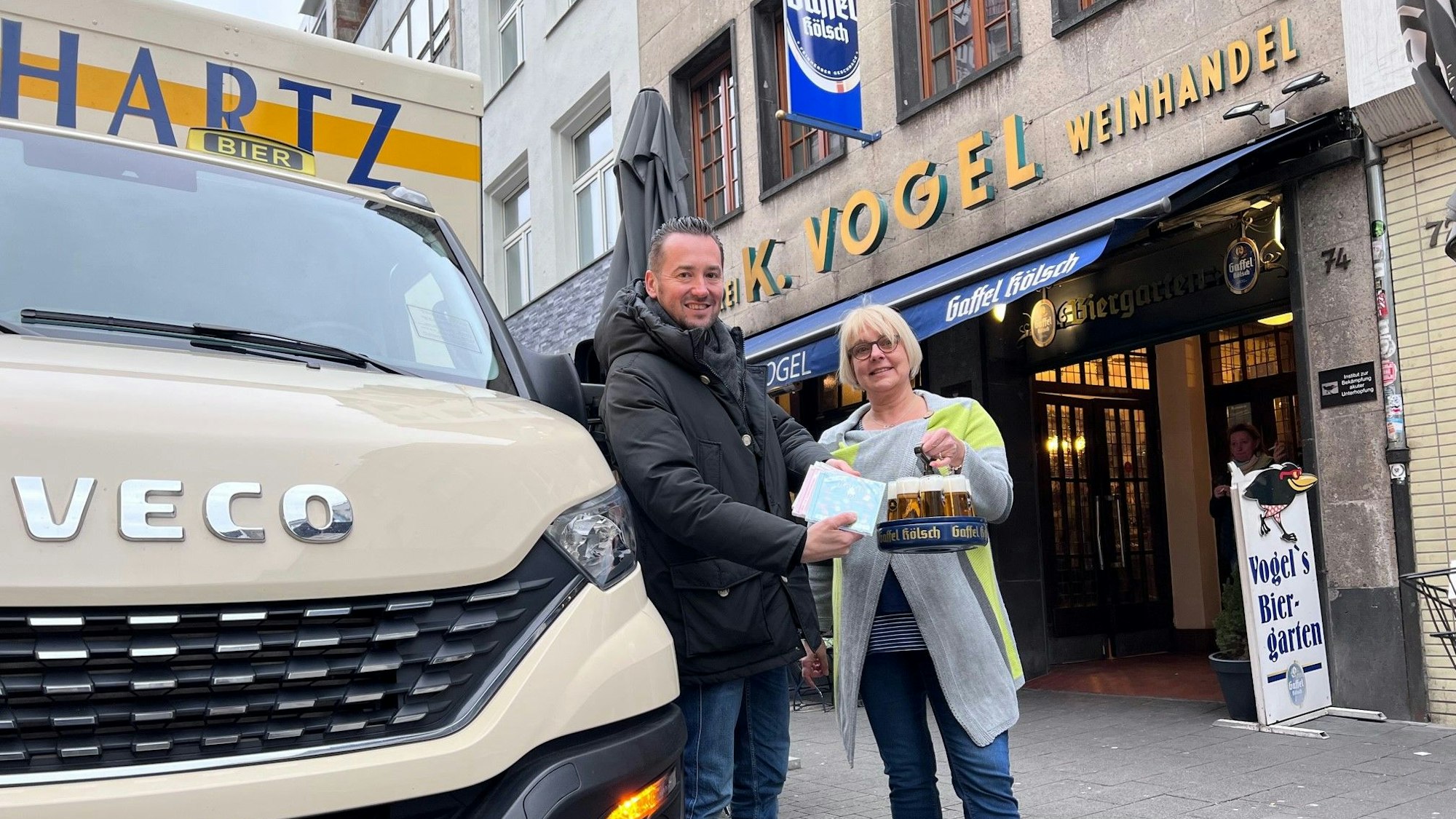 Bierlieferant Daniel Siebert (l.) übergibt am Eigelstein der Inhaberin des Weinhaus Vogel, Angelika, CDs.