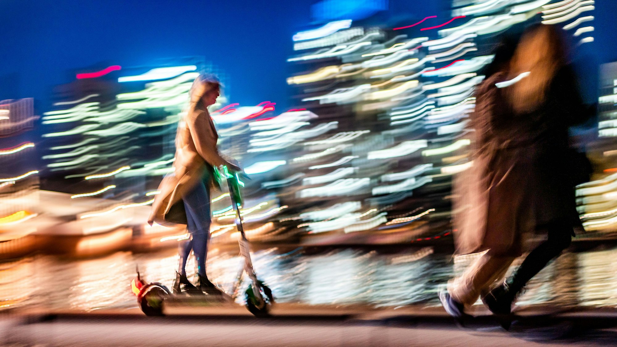 Vor den erleuchteten Hochhäusern ist am Abend eine Frau auf einem E-Scooter unterwegs. Der Wischeffekt entstand durch das Mitziehen der Kamera bei einer Belichtungszeit von einer achtel Sekunde.