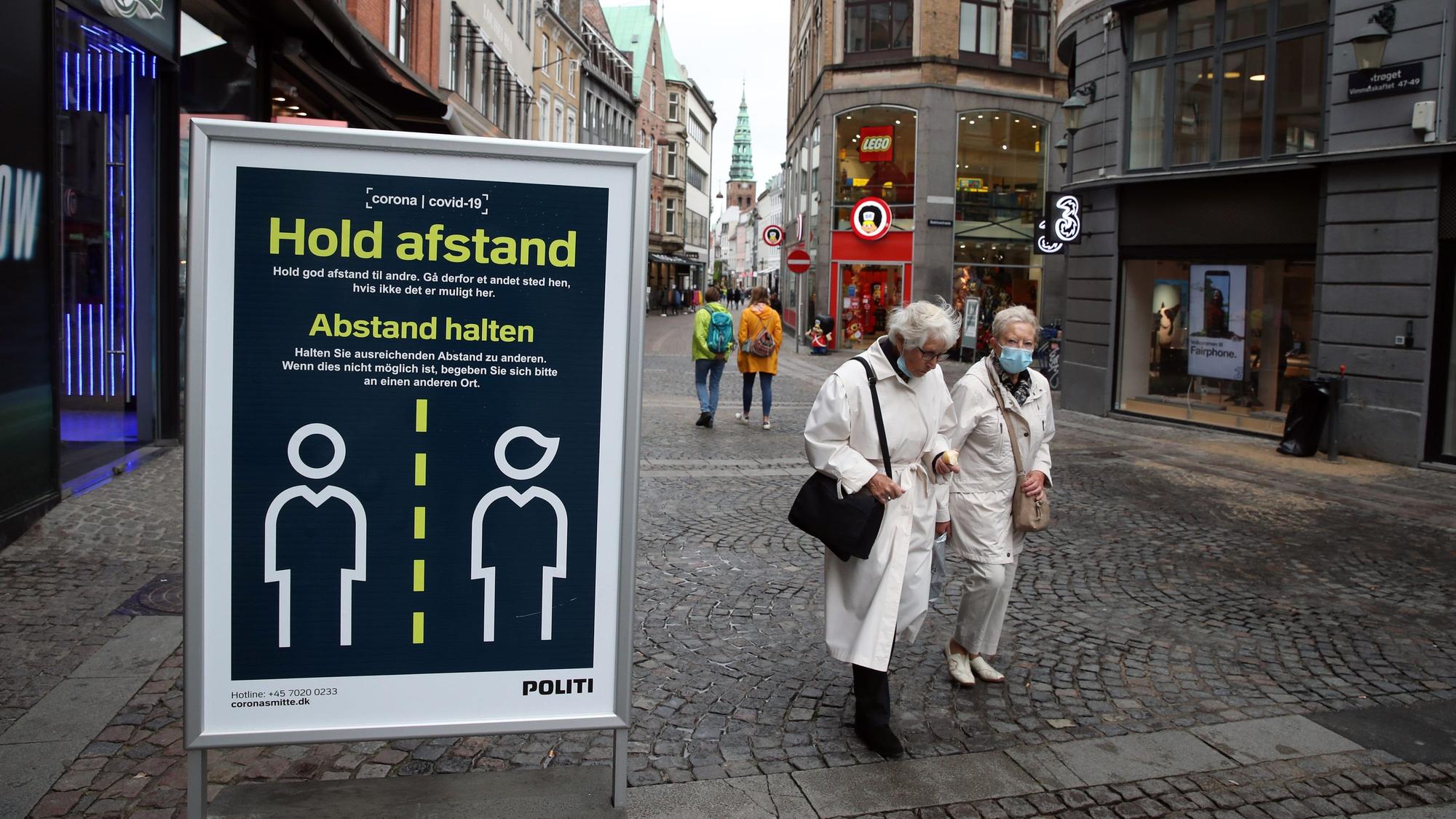 „Abstand halten“ steht auf einem Schild in einer Straße in Kopenhagen, Passanten laufen daran vorbei.
