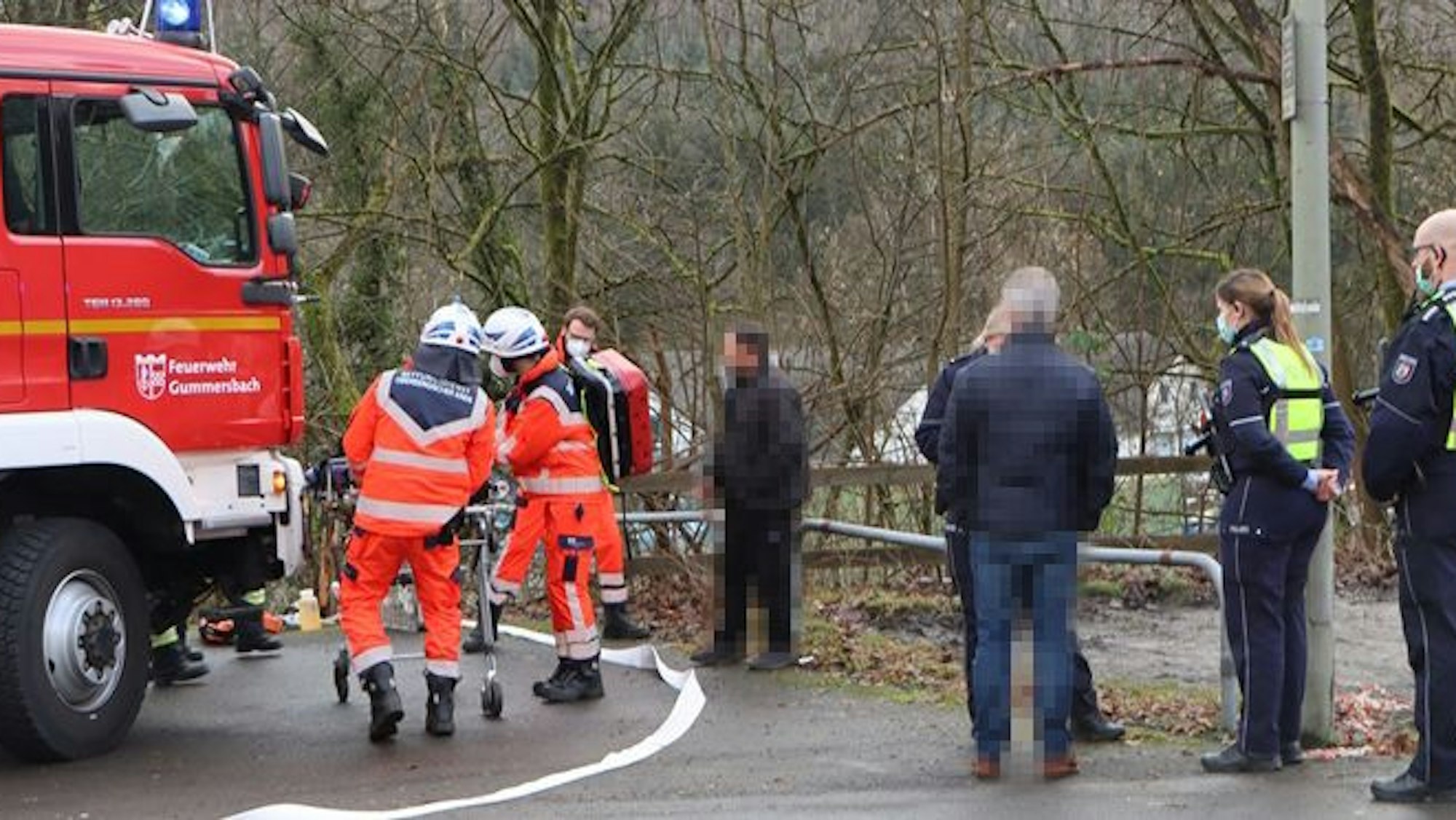 Einsatzkräfte von Polizei und Feuerwehr stehen auf einer Straße, zwei Personen schieben eine Trage.