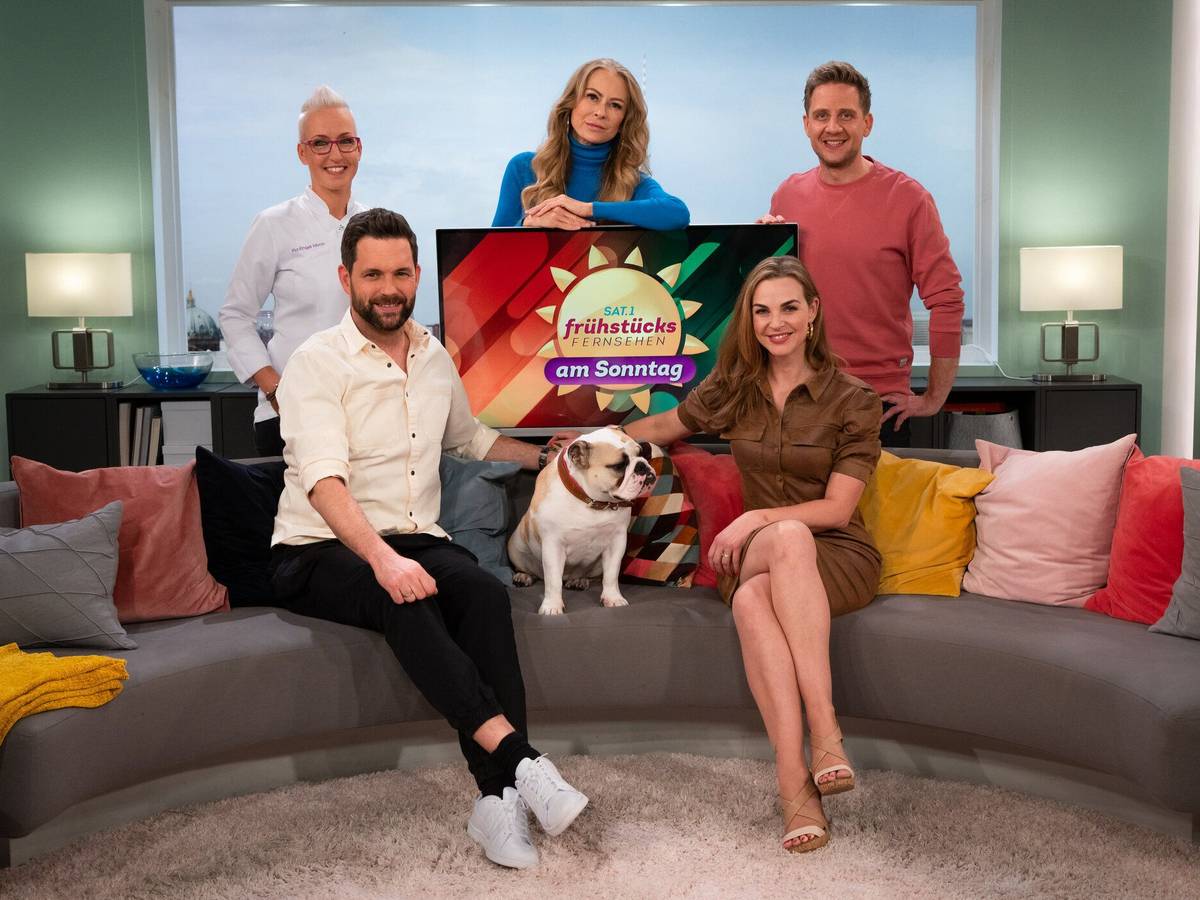 Matthias Killing und Annika Lau sitzen zusammen mit einem Hund auf dem Sofa. Ihre drei Kollegen stehen hinter dem Sofa.