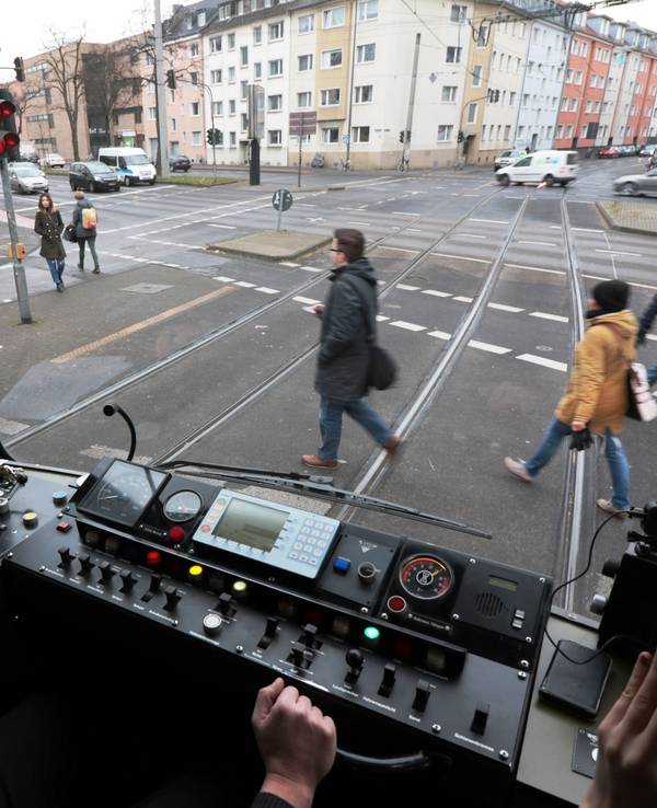 Blick aus dem Führerstand einer KVB-Bahn auf die Straße, wo Menschen die Gleise überqueren.