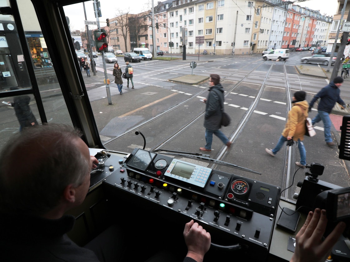 Blick aus dem Führerstand einer KVB-Bahn auf die Straße, wo Menschen die Gleise überqueren.