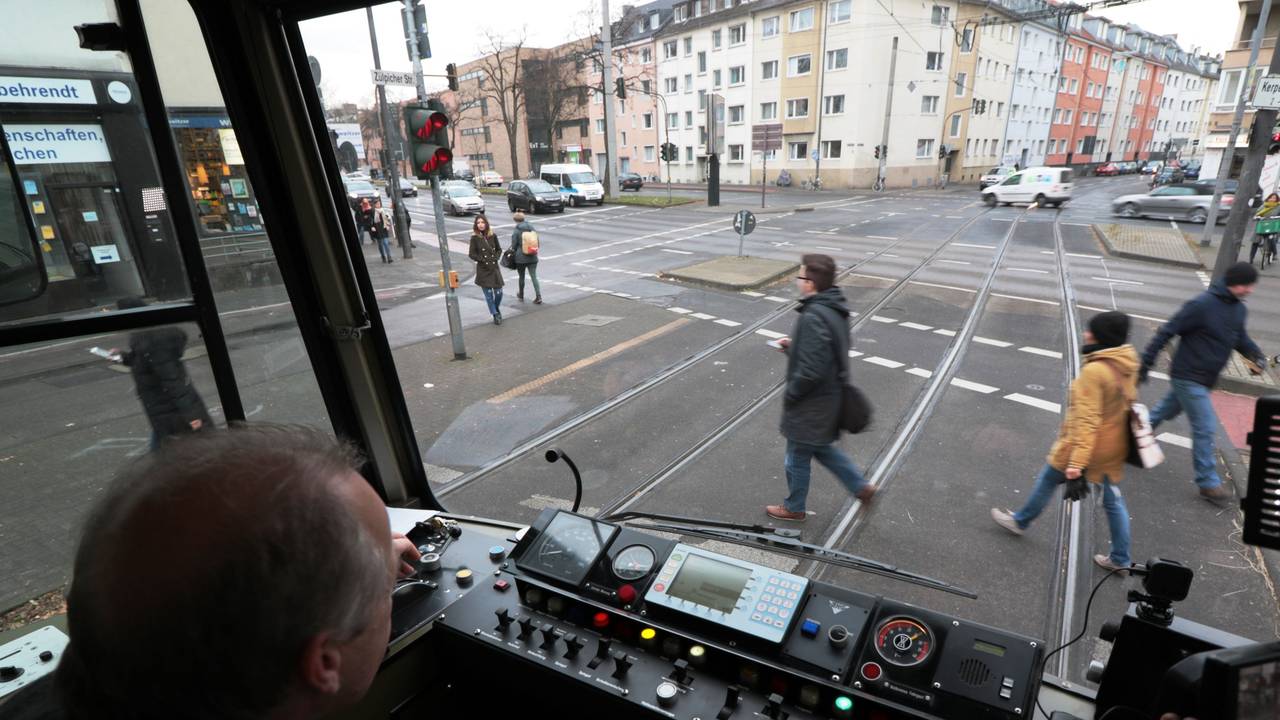 Blick aus dem Führerstand einer KVB-Bahn auf die Straße, wo Menschen die Gleise überqueren.