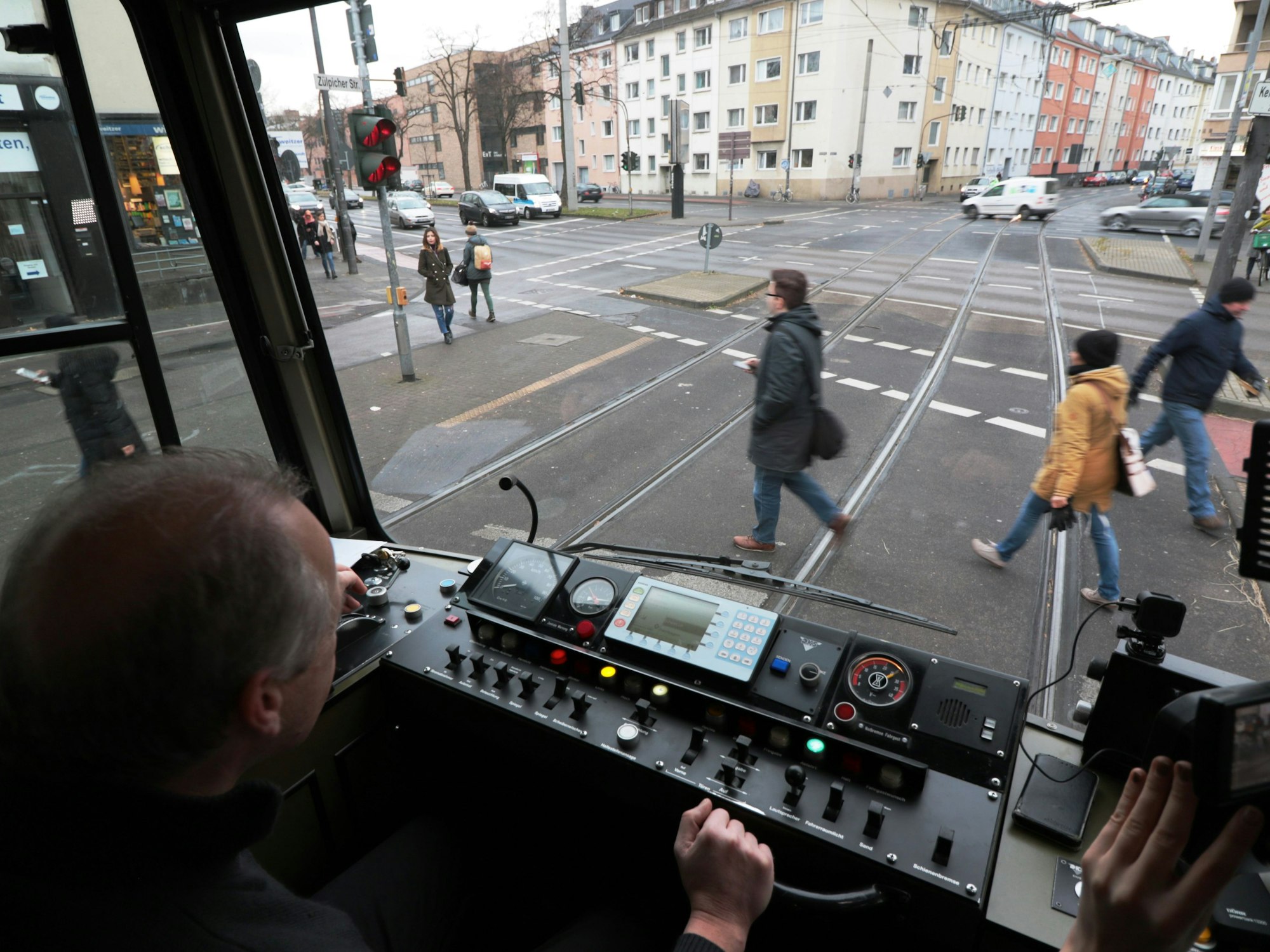 Blick aus dem Führerstand einer KVB-Bahn auf die Straße, wo Menschen die Gleise überqueren.