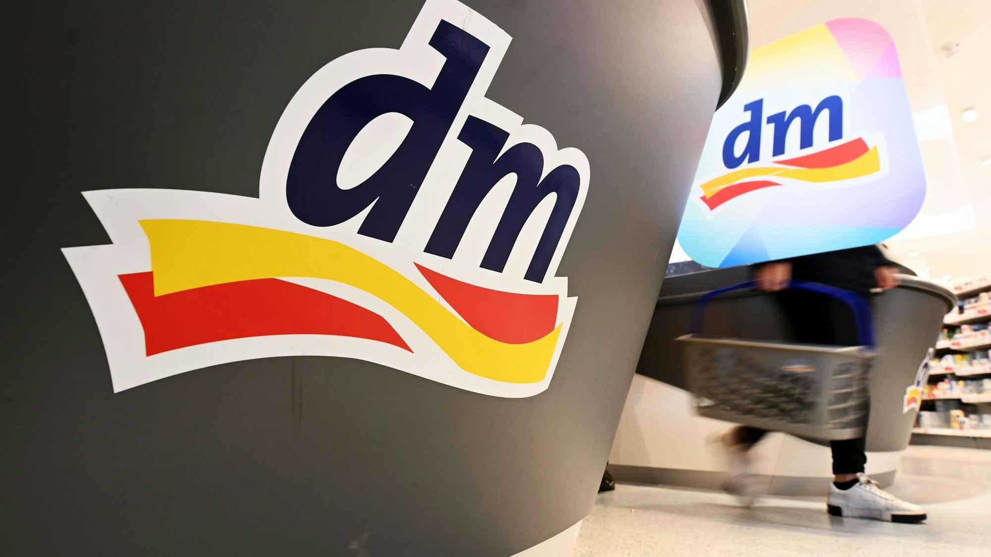 Eine Person geht in einer Filiale der Drogeriemarktkette dm mit einem Einkaufskorb am Kassenbereich vorbei.