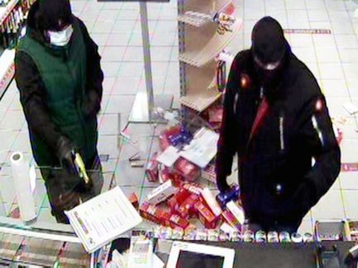 Die beiden maskierten Täter stehen vor der Kasse. Einer von ihnen hält einen Revolver.