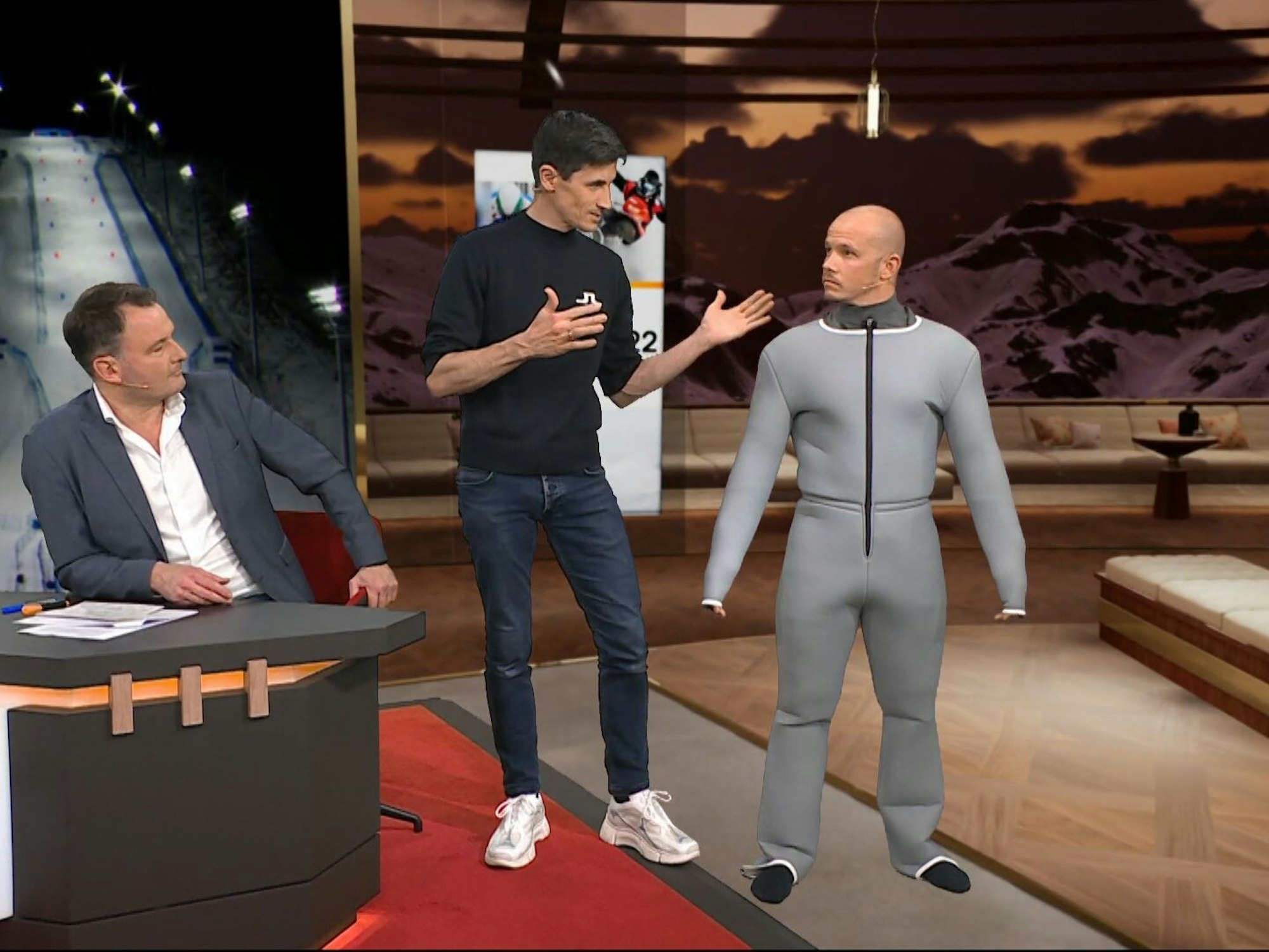 Moderator Thomas Wagner (l.) schaut zu, wie Ex-Skisprung-Star Martin Schmitt (M.) Fabian Hambüchen die Skianzug-Vermessung erklärt.