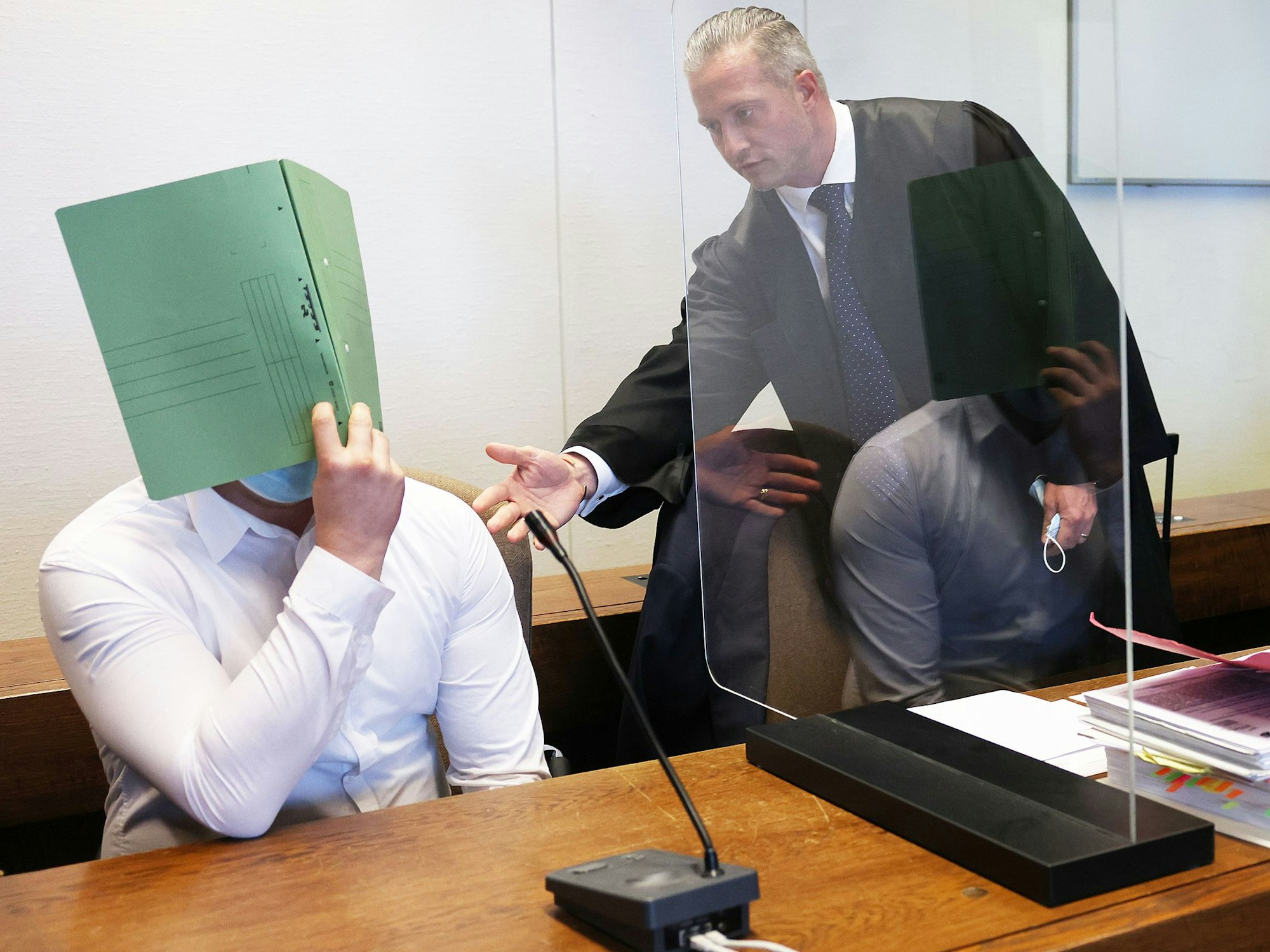 Zwei Angeklagte halten sich neben ihren Verteidigern im Gerichtssaal eine Mappe vor das Gesicht.