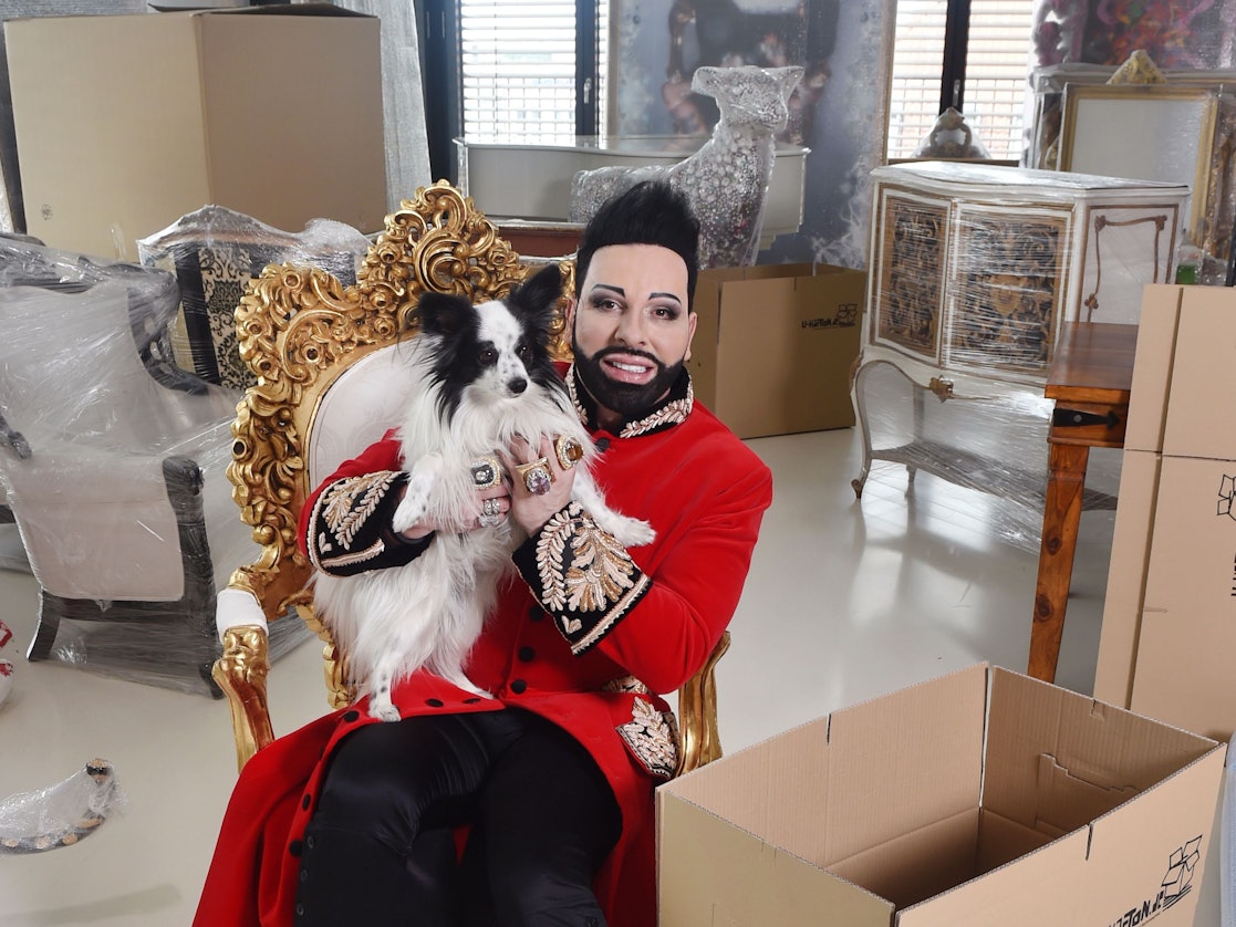 Stardesigner Harald Glööckler sitzt zwischen gepackten Kisten und eingepackten Möbelstücken und Kunstobjekten.