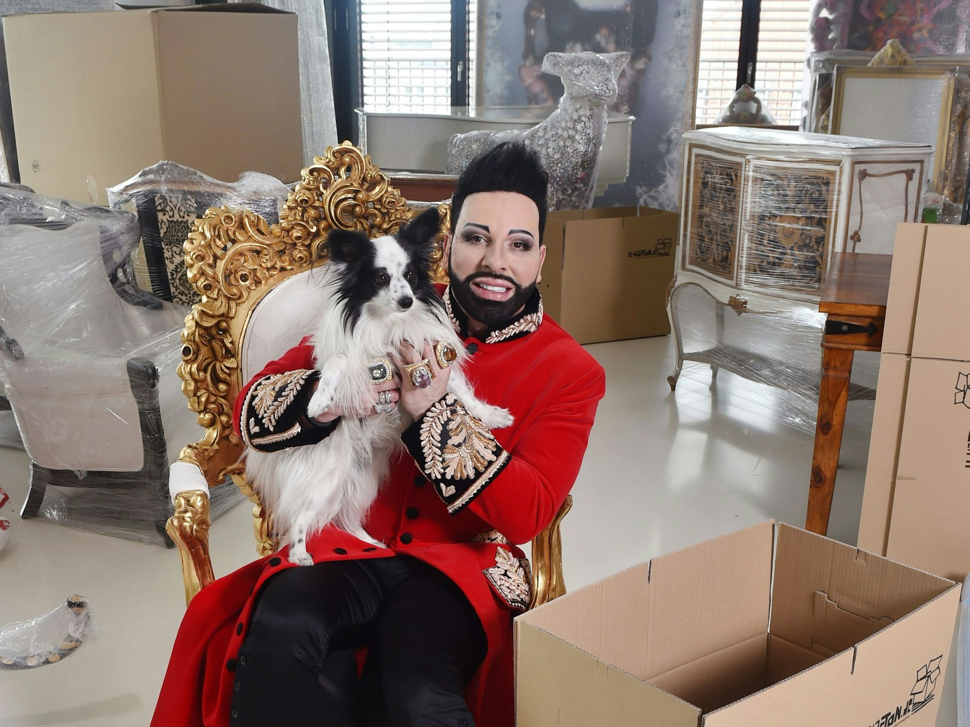 Stardesigner Harald Glööckler sitzt zwischen gepackten Kisten und eingepackten Möbelstücken und Kunstobjekten.
