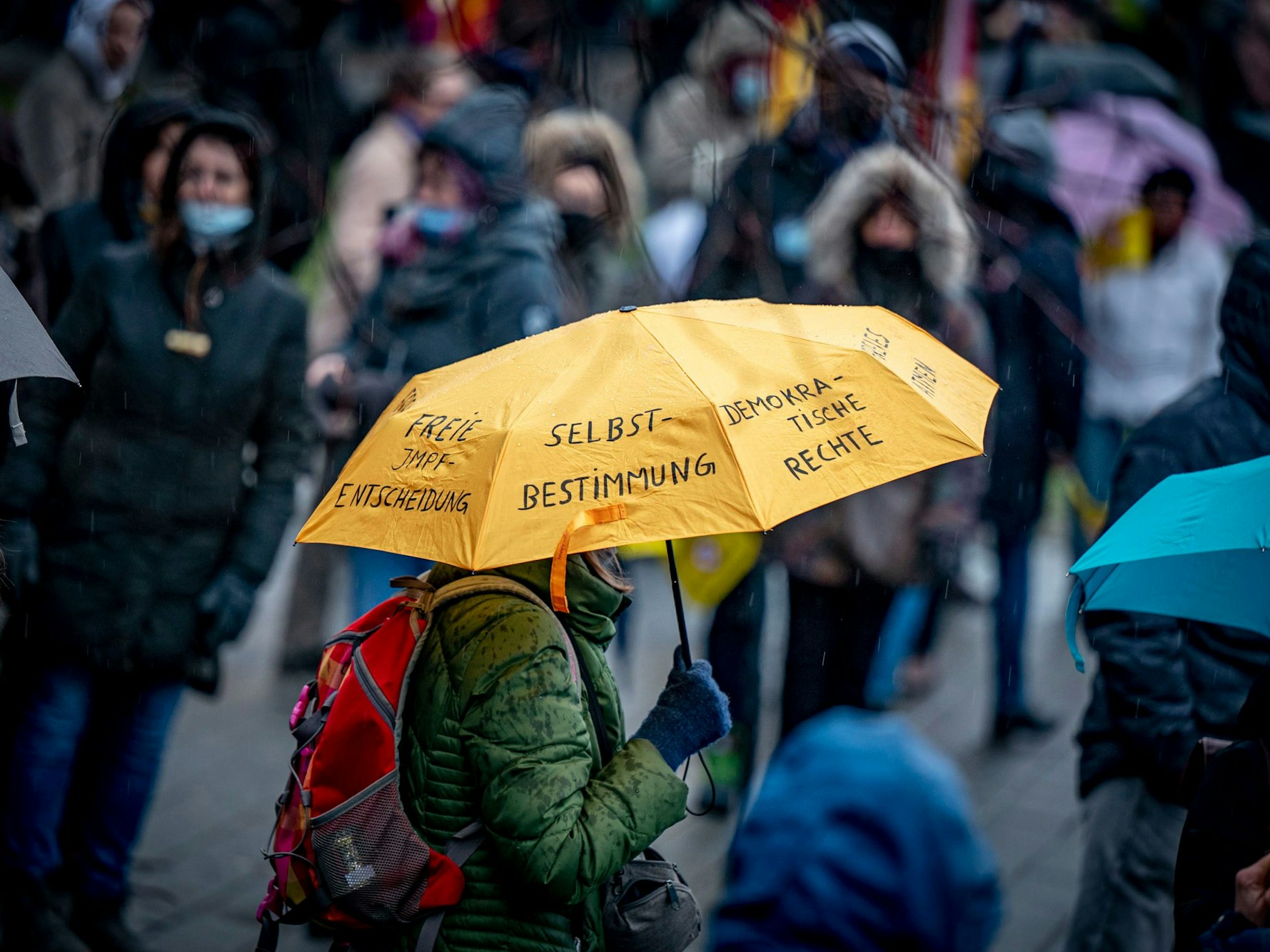 Gegner der Corona-Maßnahmen nehmen an einer Demonstration teil, eine Person hält einen Regenschirm, auf dem u. a. „Selbstbestimmung“ und „Demokratische Rechte“ steht.
