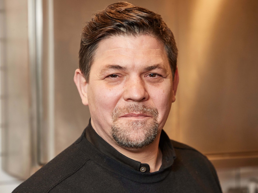 Fernsehkoch Tim Mälzer steht in der Küche seines Restaurants ««Die Gute Botschaft»». Dabei lächelt er leicht in die Kamera.