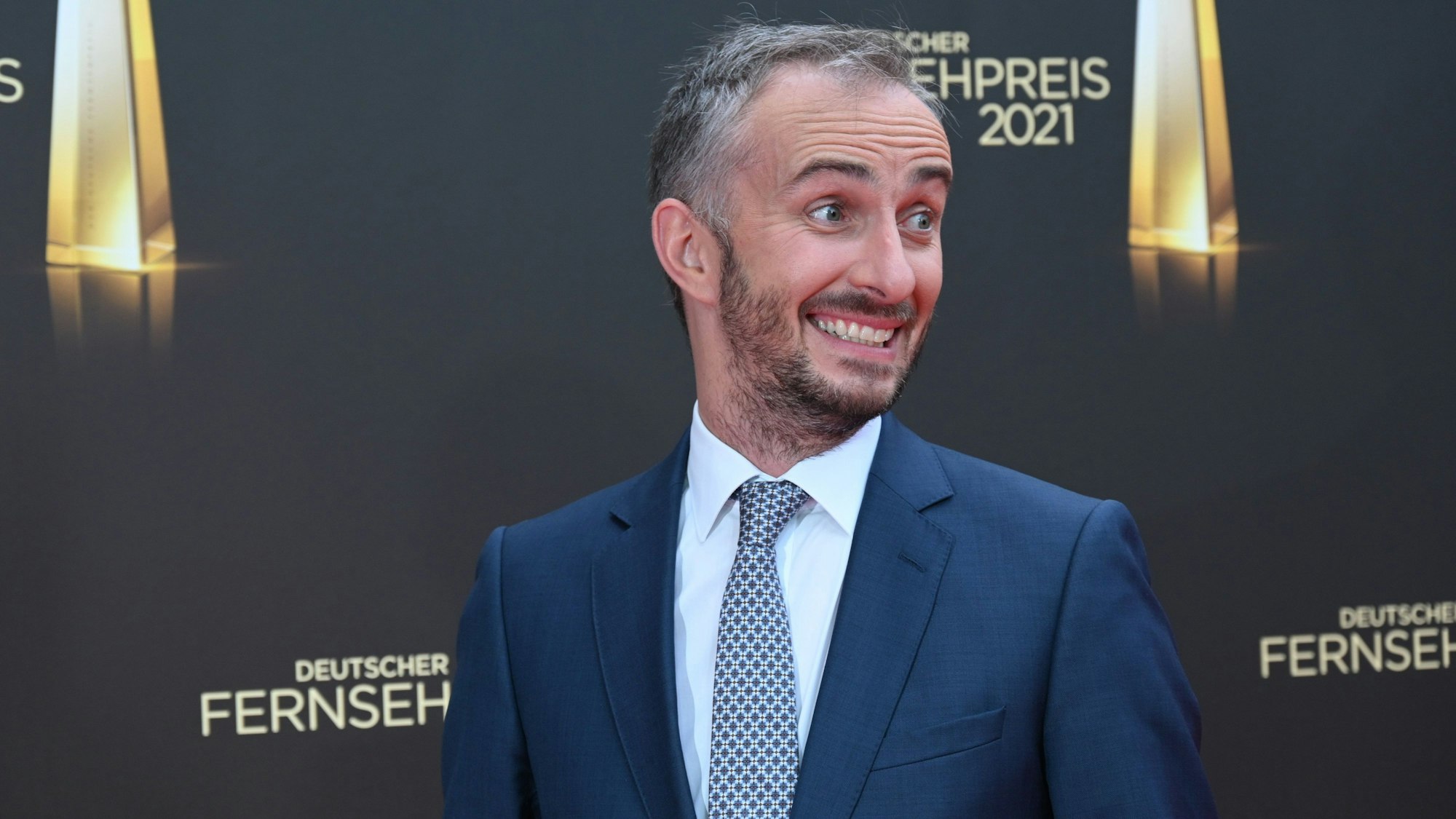 Jan Böhmermann kommt zur Verleihung des Deutschen Fernsehpreises 2021 im Tanzbrunnen. Der Satiriker steht vor einer Wand mit dem Logo der Veranstaltung und grinst schief an der Kamera vorbei.