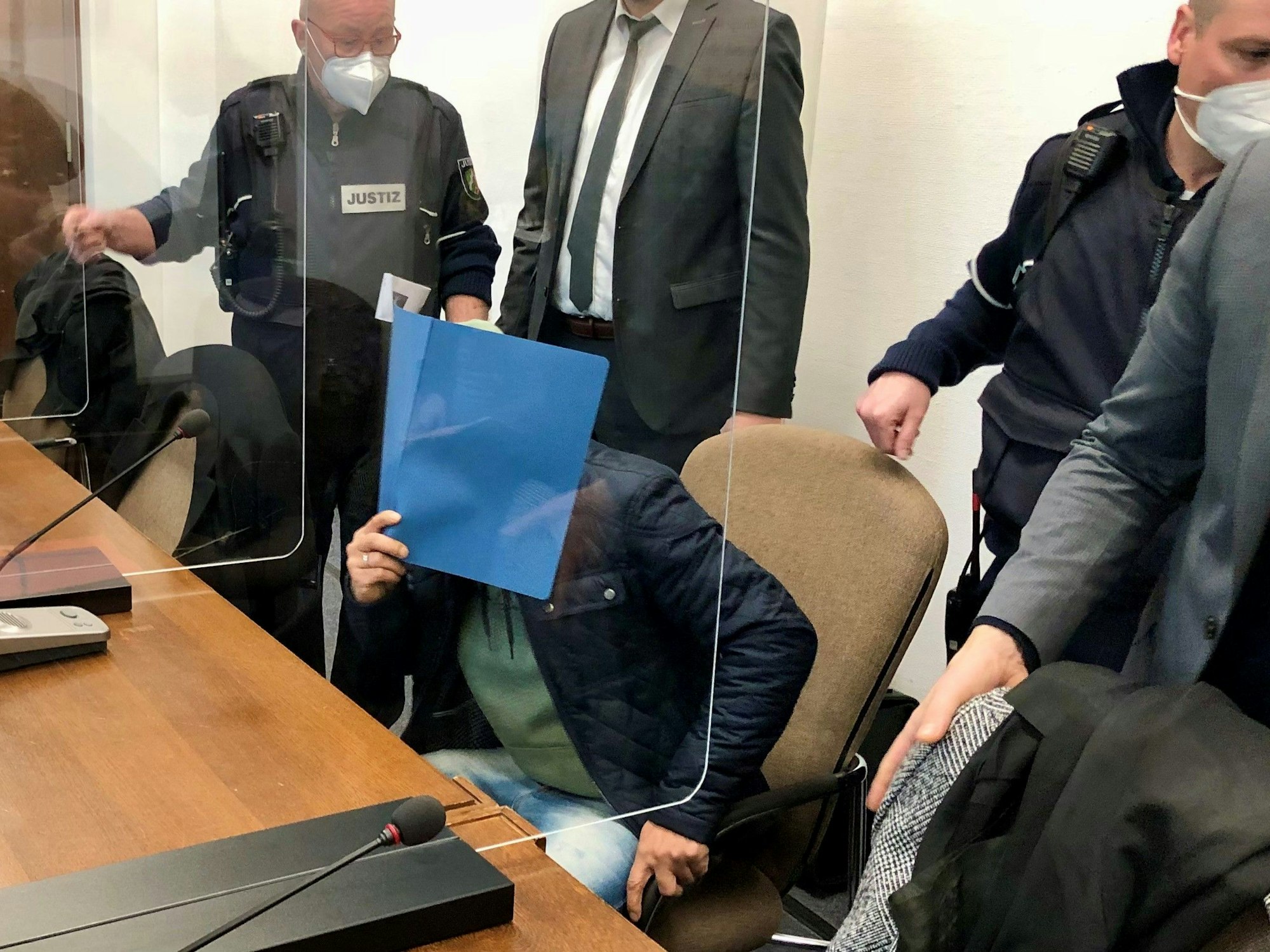 Ein Mann sitzt im Gericht auf der Anklagebank und hält sich eine Mappe vor das Gesicht.