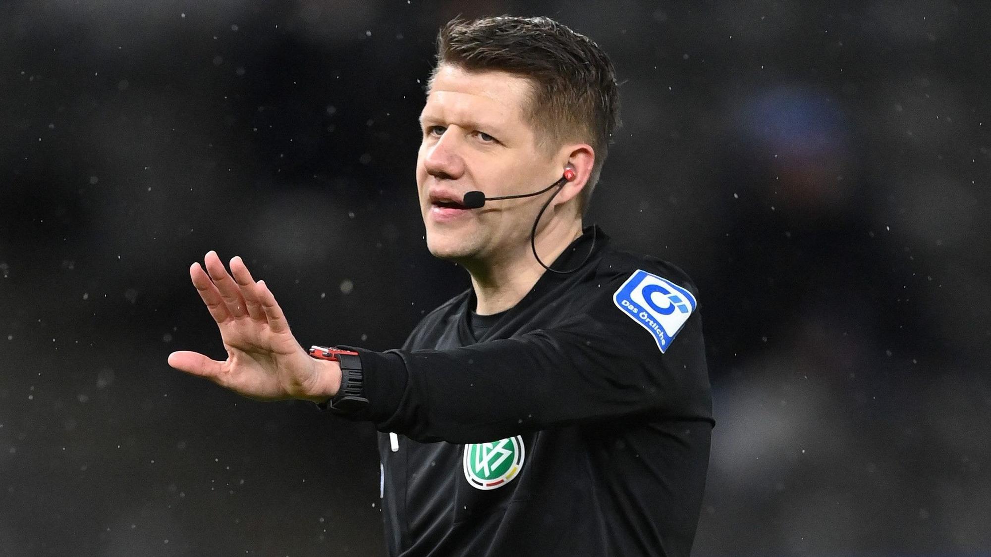 Schiedsrichter Patrick Ittrich hält protestierende Spieler mit ausgefahrenem Arm zurück.