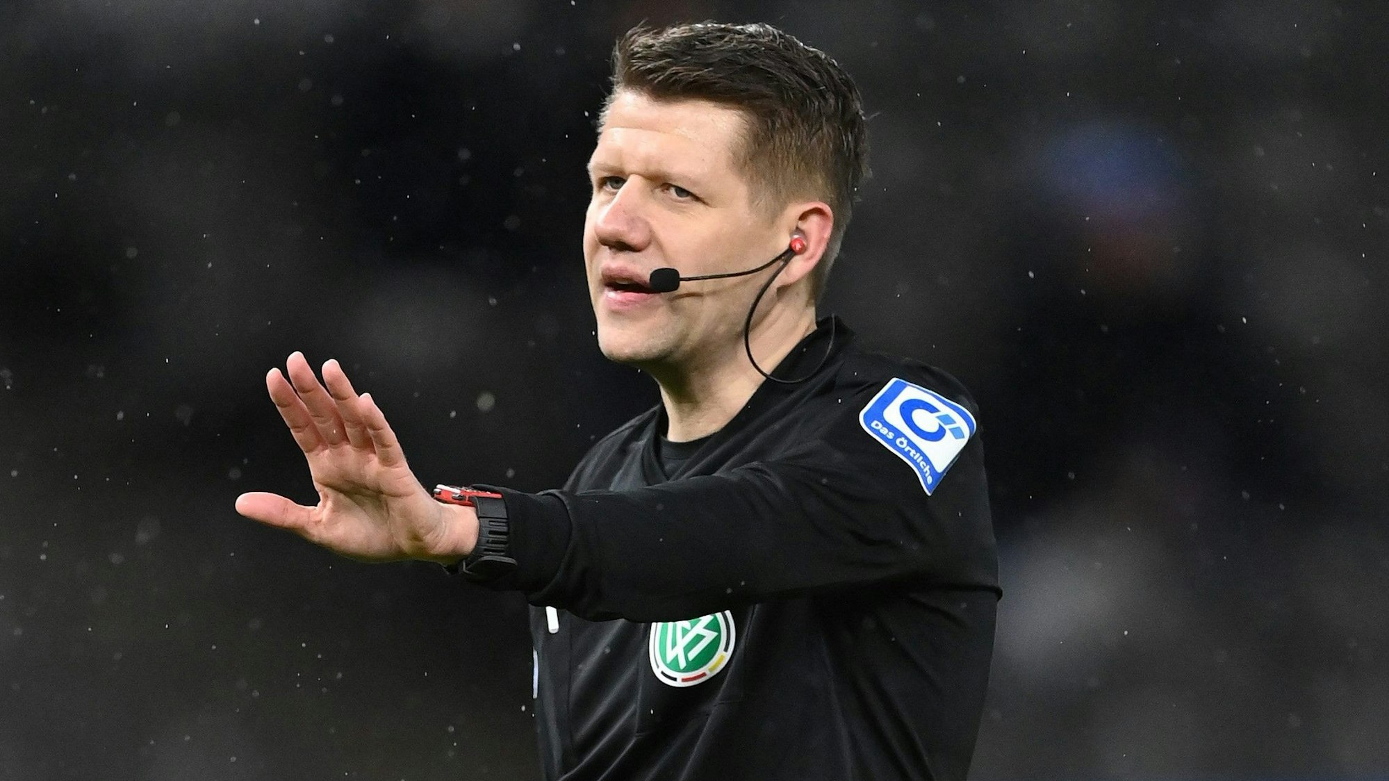 Schiedsrichter Patrick Ittrich hält protestierende Spieler mit ausgefahrenem Arm zurück.