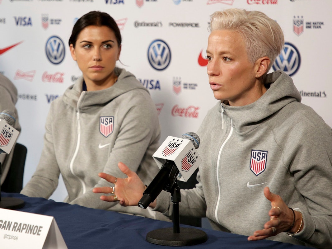 Frauen-Nationalmannschaft der USA bei einer Pressekonferenz. Megan Rapinoe spricht ins Mikrofon. Team-Kollegin Alex Morgan schaut sie an.