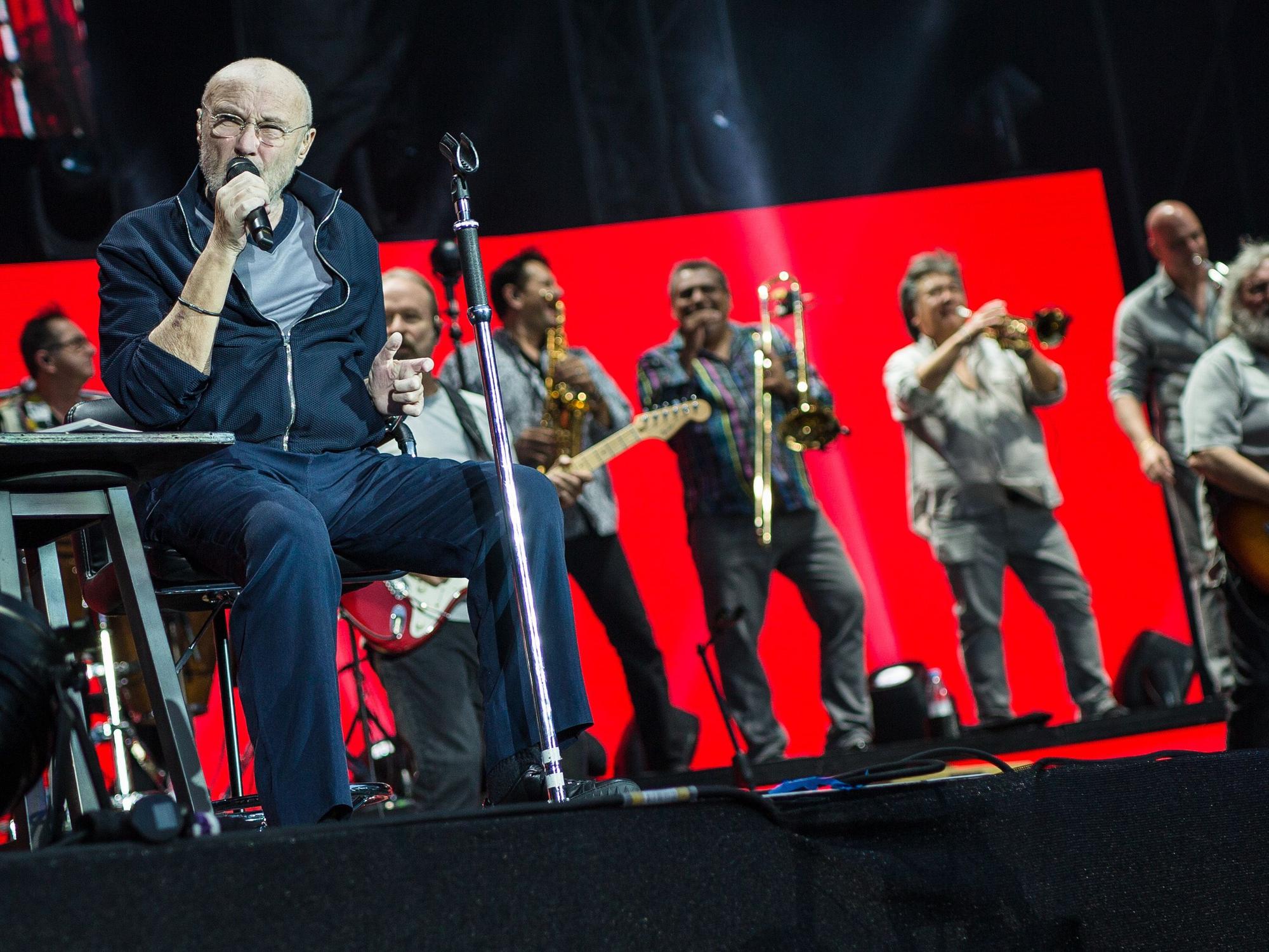 Sänger Phil Collins sitzt auf der Bühne und singt, hinter ihm stehen die Bandmitglieder.