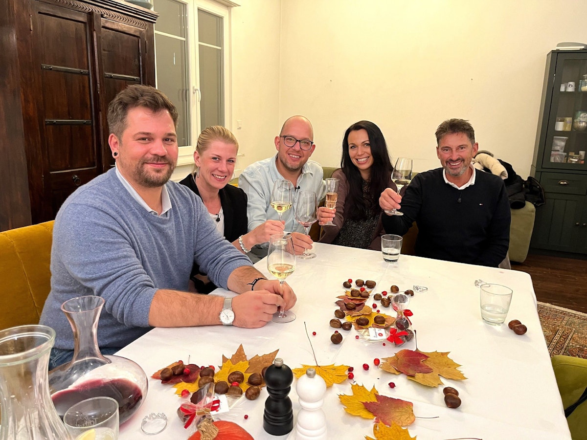 Die Das Perfekte Dinner-Kandidaten Marcus, Simone, Gastgeber Andreas, Sabrina und Jürgen sitzen an Andreas Tisch und lächeln.