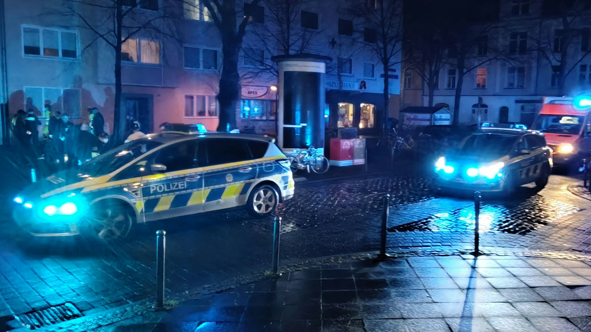 Streifenwagen sowie ein RTW stehen auf einer Straße, mehrere Polizisten befinden sich vor einem Haus.