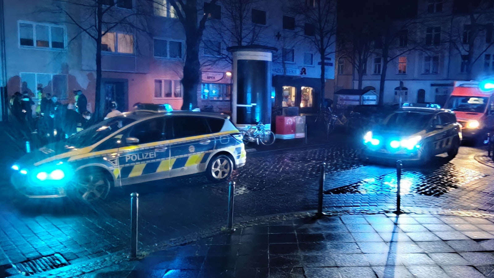 Streifenwagen sowie ein RTW stehen auf einer Straße, mehrere Polizisten befinden sich vor einem Haus.