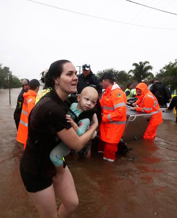 28.02.2022, Australien, Lismore: Eine Frau trägt ein Baby, während Menschen mit kleinen Booten durch das Hochwasser fahren. Sintflutartiger Regen hat die schweren Überschwemmungen im Osten Australiens weiter verschärft.