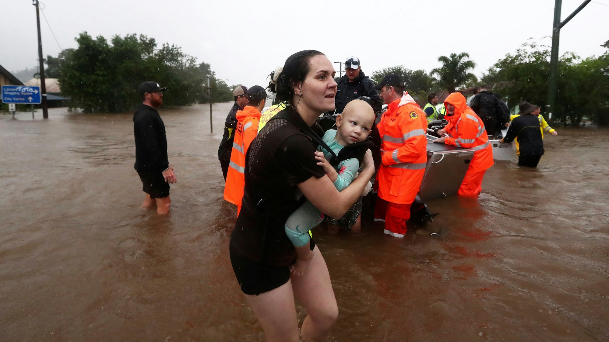 28.02.2022, Australien, Lismore: Eine Frau trägt ein Baby, während Menschen mit kleinen Booten durch das Hochwasser fahren. Sintflutartiger Regen hat die schweren Überschwemmungen im Osten Australiens weiter verschärft.