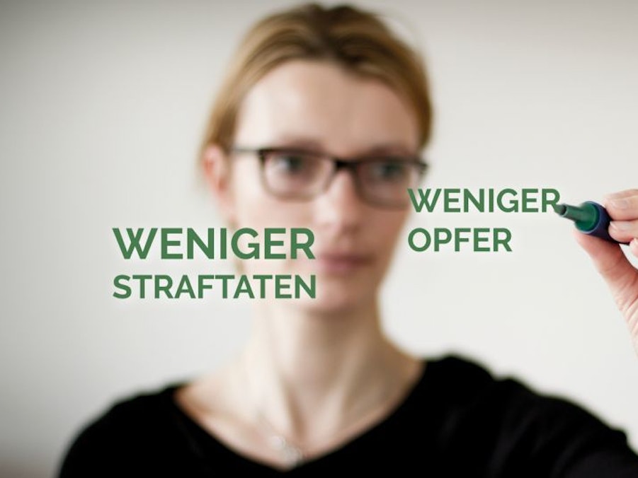 Eine Frau schreibt mit einem Stift auf eine Glasscheibe, auf der die Begriffe „Weniger Straftaten“ und „Weniger Opfer“ zu sehen sind.