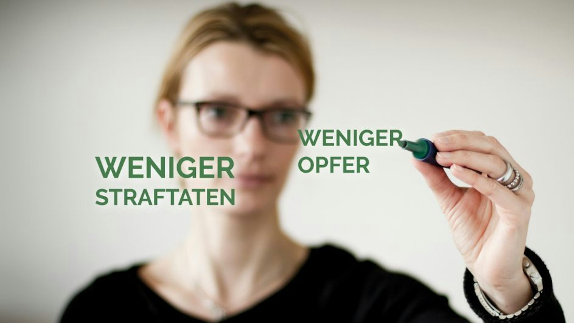 Eine Frau schreibt mit einem Stift auf eine Glasscheibe, auf der die Begriffe „Weniger Straftaten“ und „Weniger Opfer“ zu sehen sind.