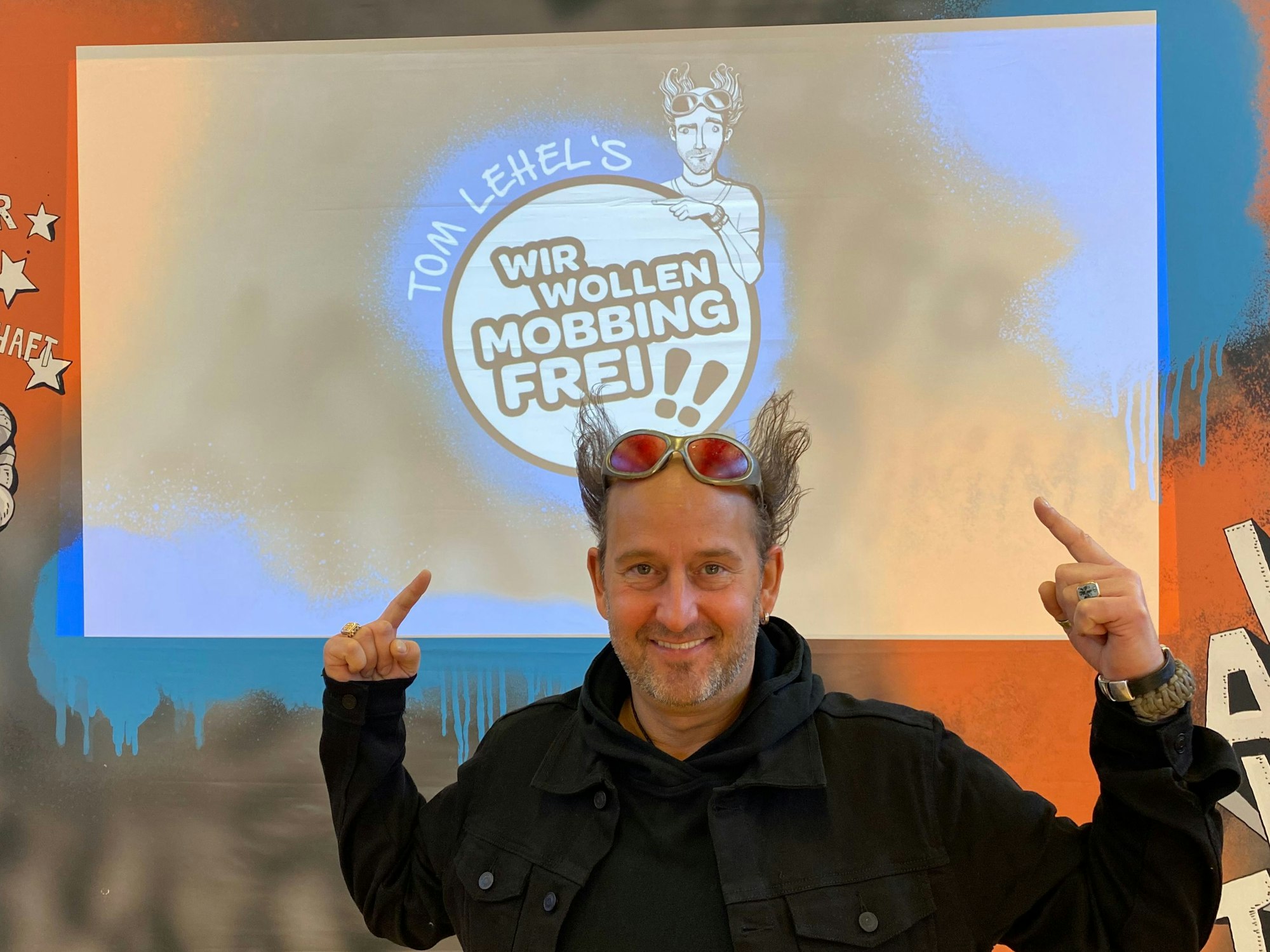 Tom Lehel zeigt in einer Kölner Grundschule auf das Logo seines Programms „Tom Lehel’s WIR WOLLEN MOBBINGFREI!!“