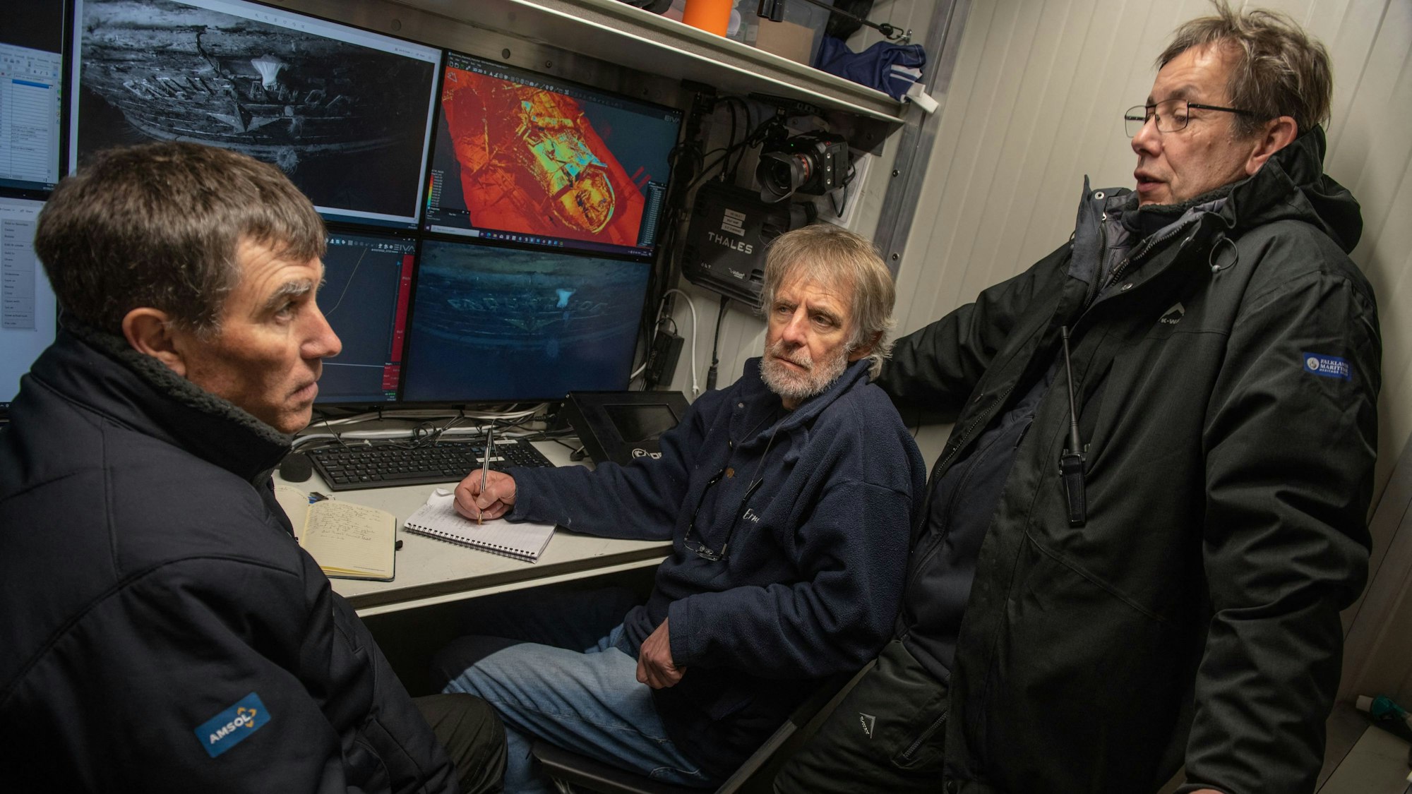 Zu sehen sind die Mitglieder des Expeditionsteams, welche vor vier Monitoren mit Bildern des Schiffes sitzen.