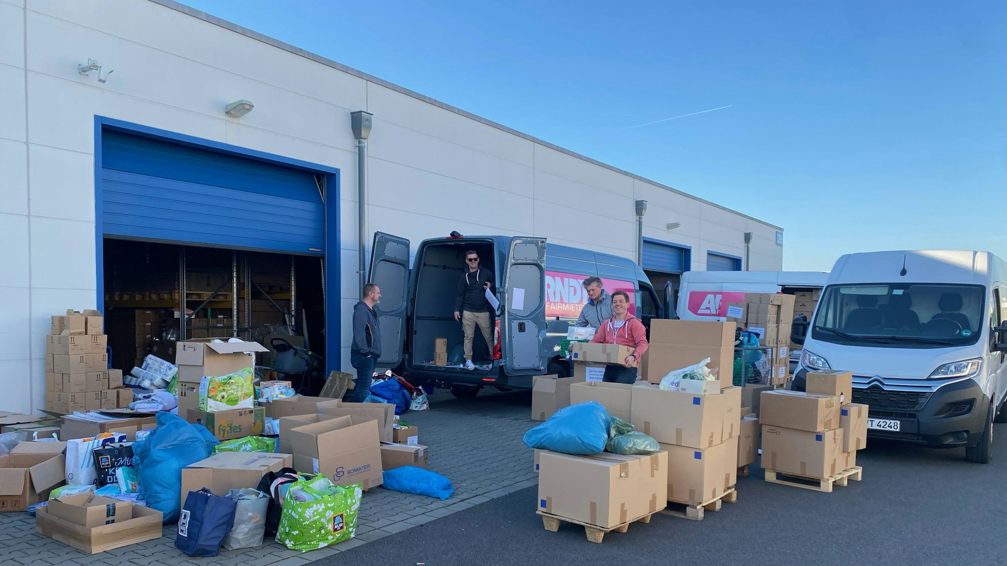 Junge Kölner vor einer Lagerhalle, die mit Umzugskartons voll mit Sachspenden gefüllt ist