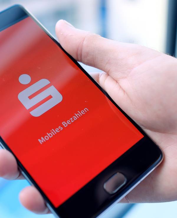 Ein Mann hält ein Mobiltelefon, auf dem der Startbildschirm der Mobiles-Bezahlen-App der Sparkasse dargestellt wird, in der Hand.