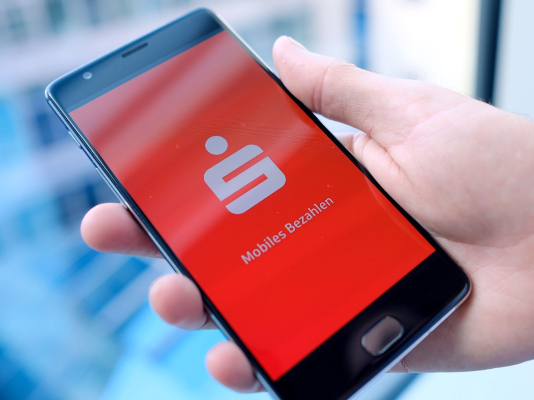 Ein Mann hält ein Mobiltelefon, auf dem der Startbildschirm der Mobiles-Bezahlen-App der Sparkasse dargestellt wird, in der Hand.