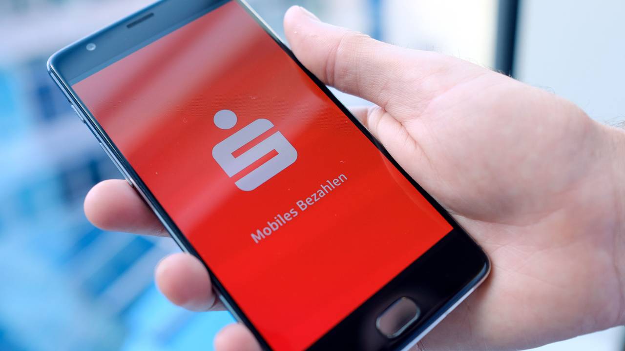Ein Mann hält ein Mobiltelefon, auf dem der Startbildschirm der Mobiles-Bezahlen-App der Sparkasse dargestellt wird, in der Hand.