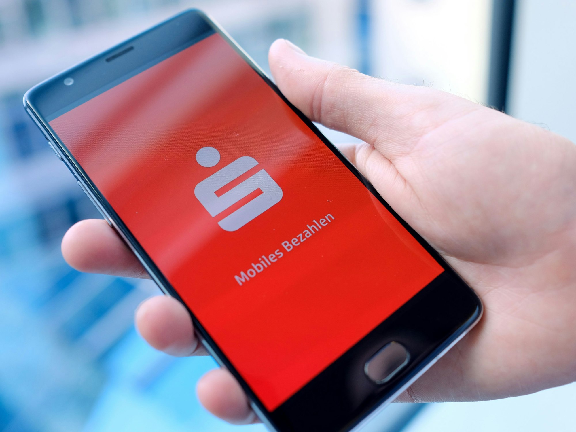 Ein Mann hält ein Mobiltelefon, auf dem der Startbildschirm der Mobiles-Bezahlen-App der Sparkasse dargestellt wird, in der Hand.