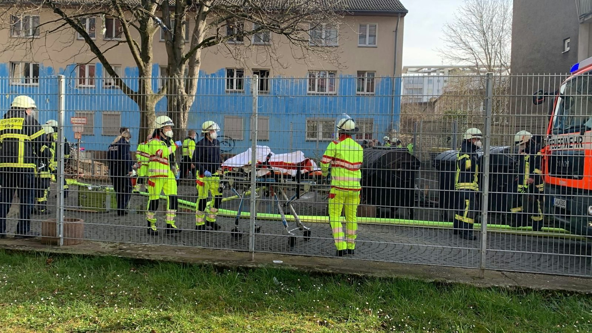 Mehrerer Feuerwehrmänner sowie eine Rettungstrage auf Rollen stehen hinter einem Zaun, auf dem Boden ist ein Löschschlauch ausgerollt.