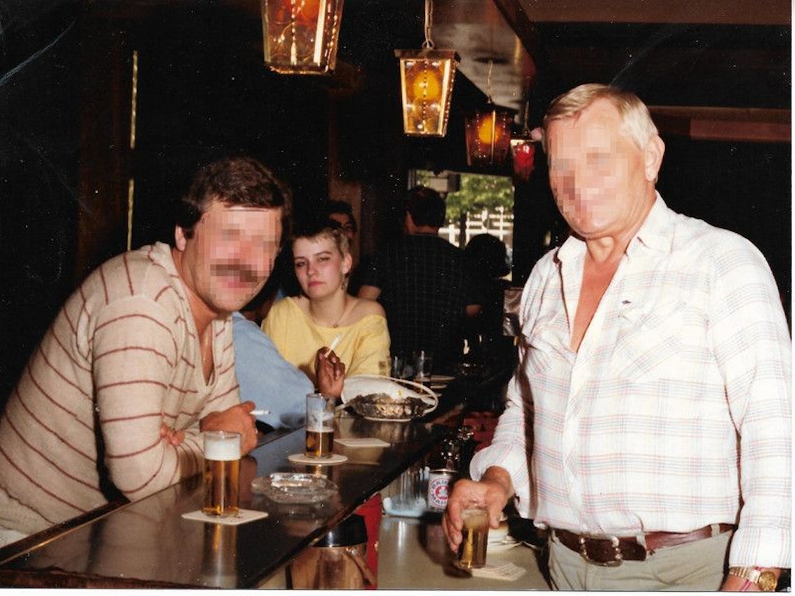 Eine junge Frau und ein Mann mit Schnäuzer sitzen an einem Kneipentresen, dahinter steht ebenfalls ein Mann. Die Gesichter der beiden Männer sind unkenntlich gemacht.