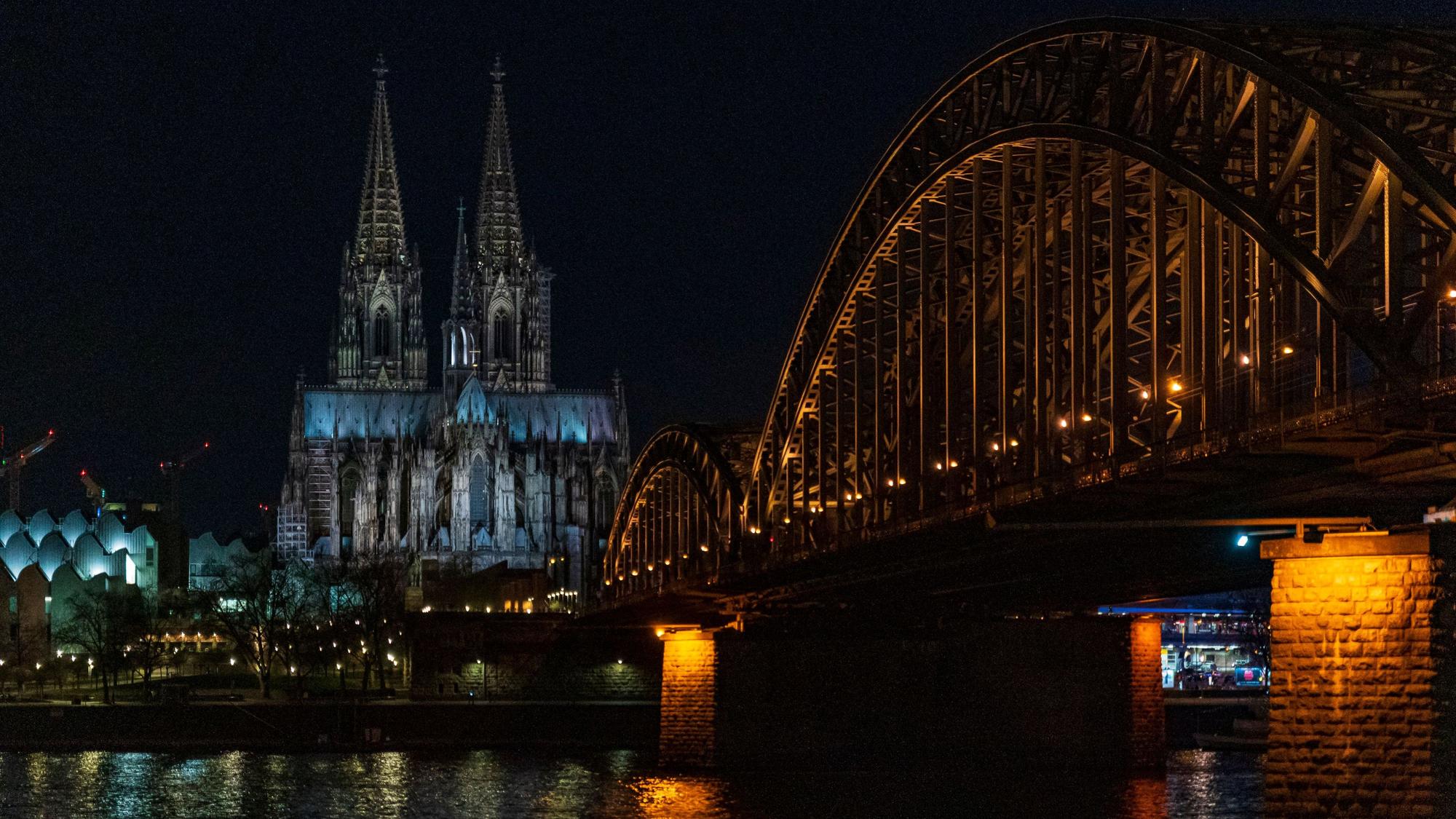 Der Kölner Dom liegt im Dunkeln, auch eine Rheinbrücke im Vordergrund ist nicht hell beleuchtet.