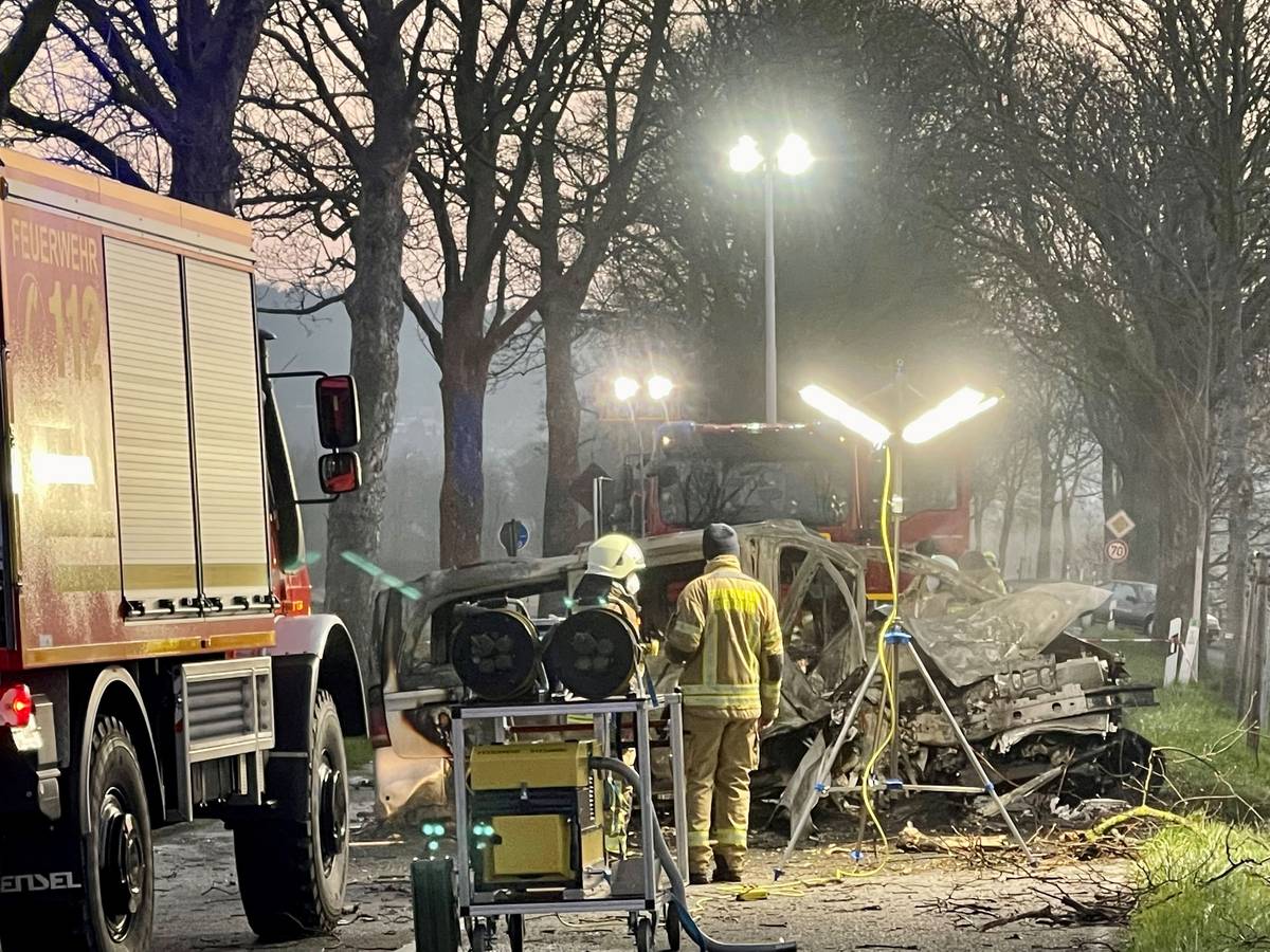 Zwei Feuerwehrmänner stehen vor dem ausgebrannten Wrack eines Transporters, in dem zwei junge Männer (beide 17) aus Siegburg gestorben sind.