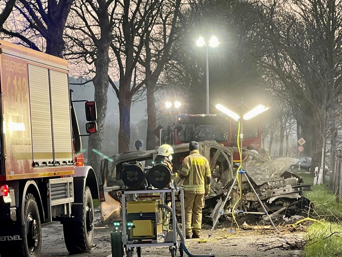 Zwei Feuerwehrmänner stehen vor dem ausgebrannten Wrack eines Transporters, in dem zwei junge Männer (beide 17) aus Siegburg gestorben sind.