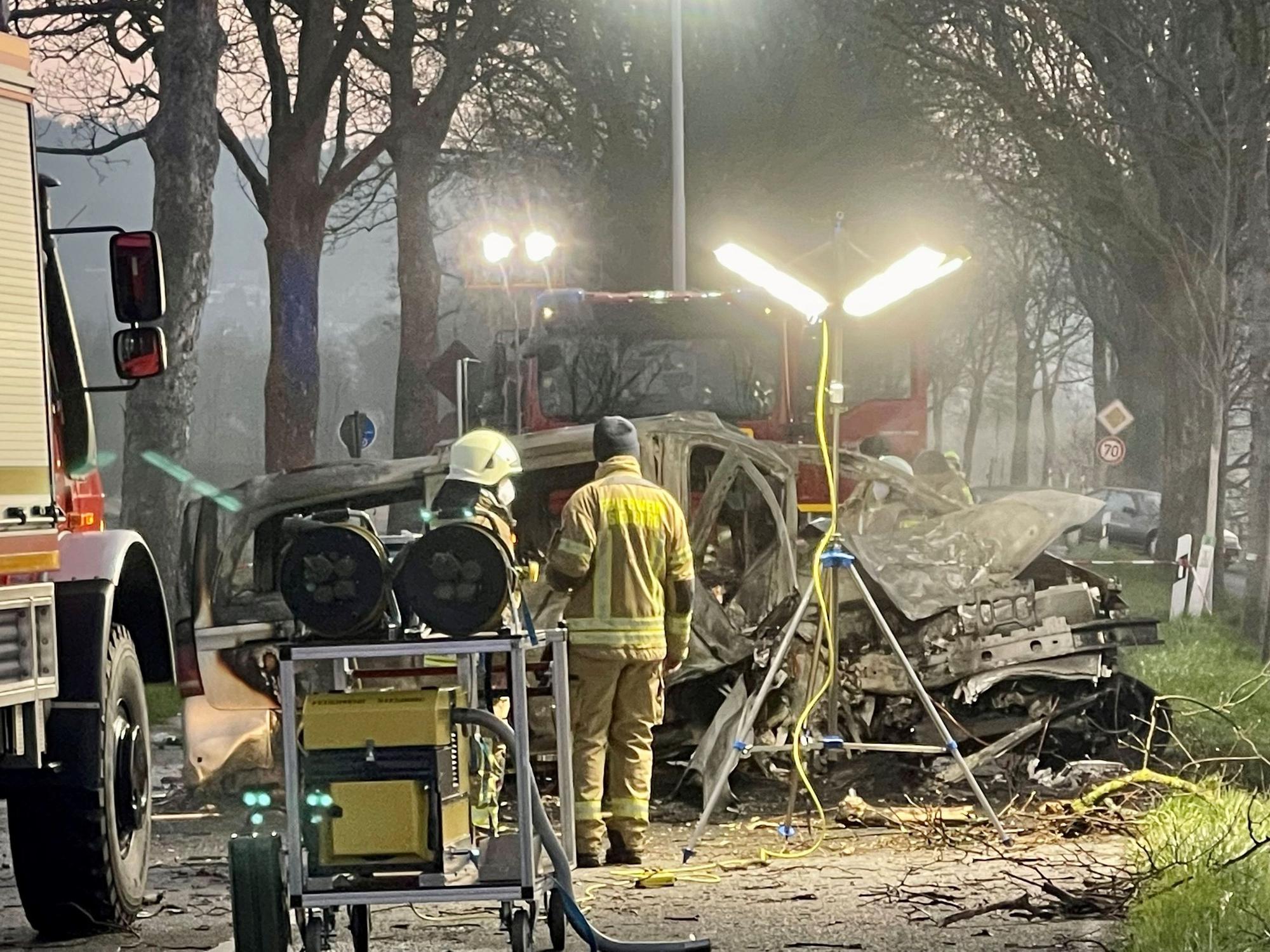 Zwei Feuerwehrmänner stehen vor dem ausgebrannten Wrack eines Transporters, in dem zwei junge Männer (beide 17) aus Siegburg gestorben sind.