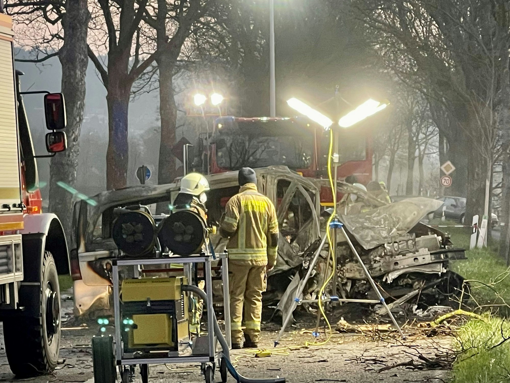 Zwei Feuerwehrmänner stehen vor dem ausgebrannten Wrack eines Transporters, in dem zwei junge Männer (beide 17) aus Siegburg gestorben sind.