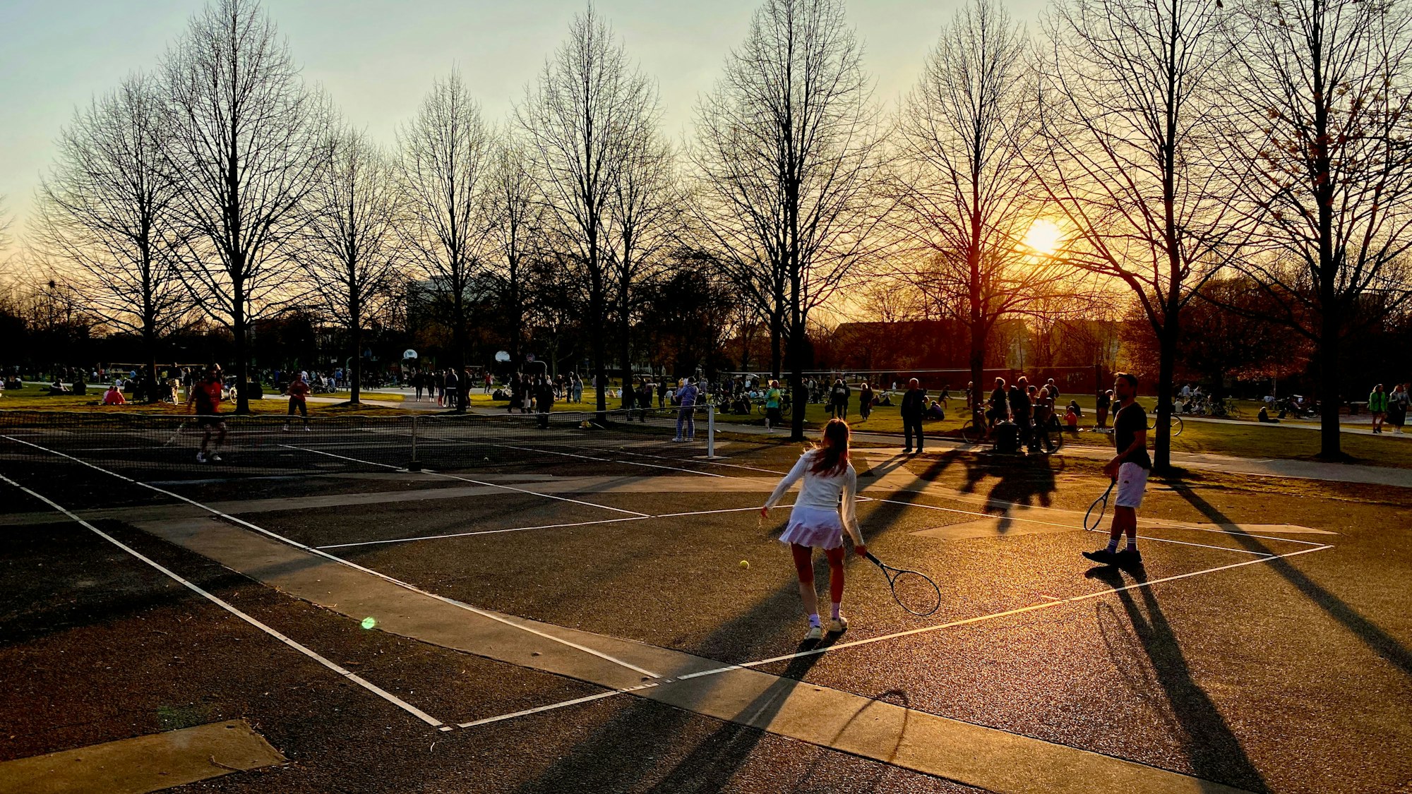 Menschen spielen in der Abendsonne auf einem öffentlichen Tennisplatz.