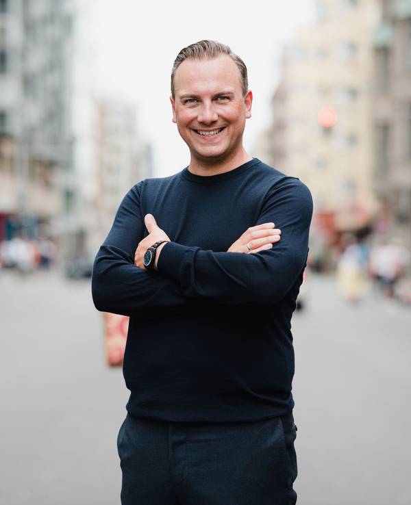 Tim Raue steht mit verschränkten Armen im Bild und lacht in die Kamera.