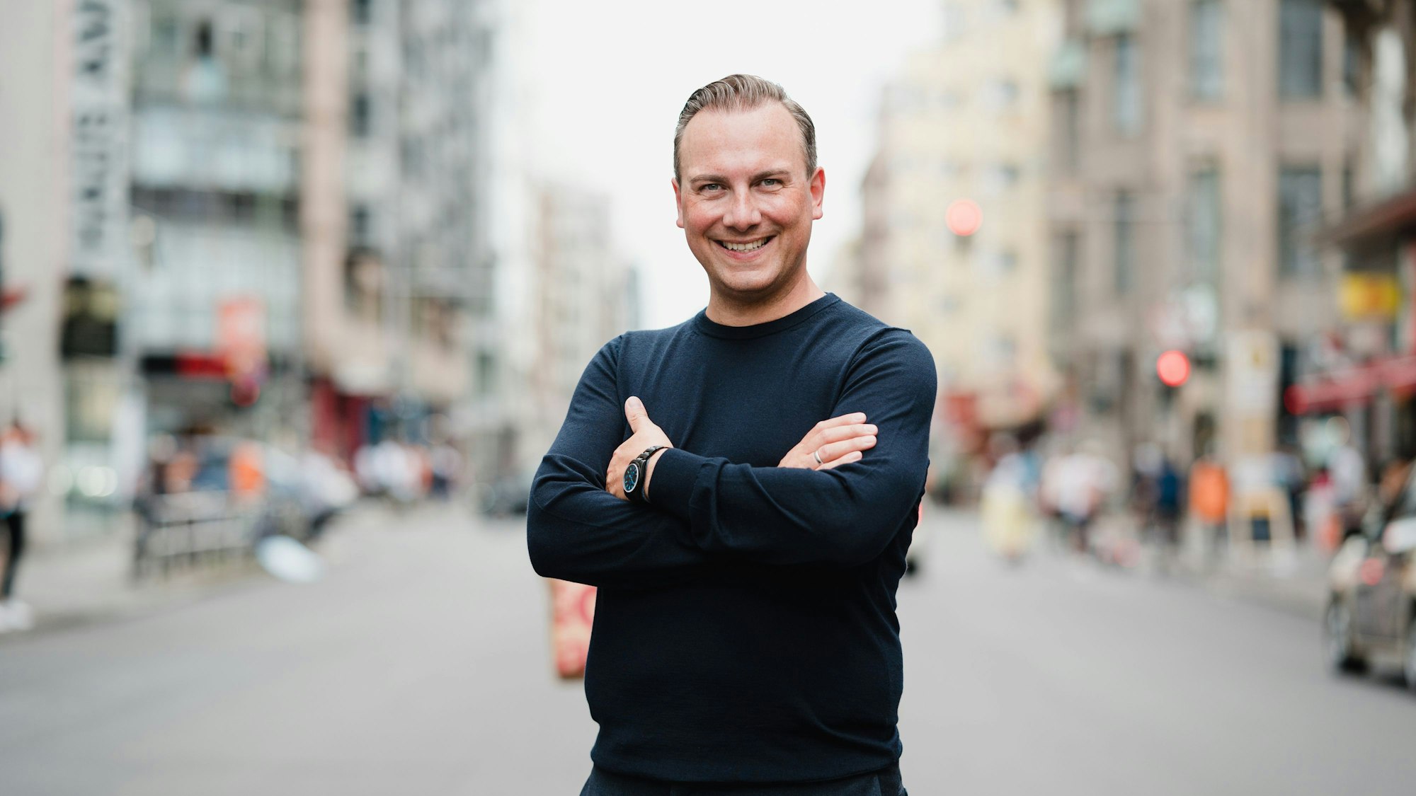 Tim Raue steht mit verschränkten Armen im Bild und lacht in die Kamera.