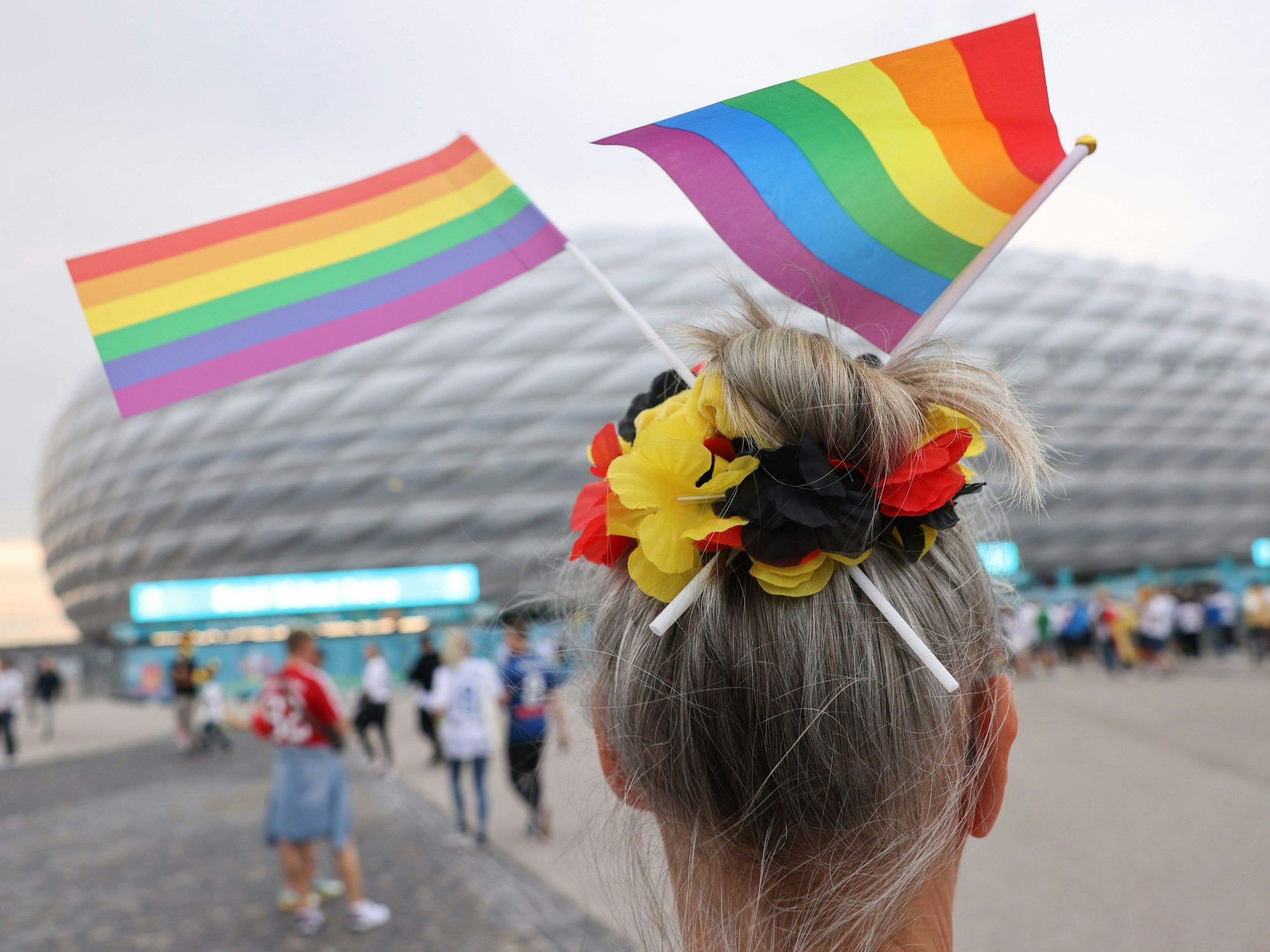 Eine Fußball-Anhängerin mit zwei kleinen Regenbogenfahnen im Haar steht vor der Münchner Allianz-Arena.