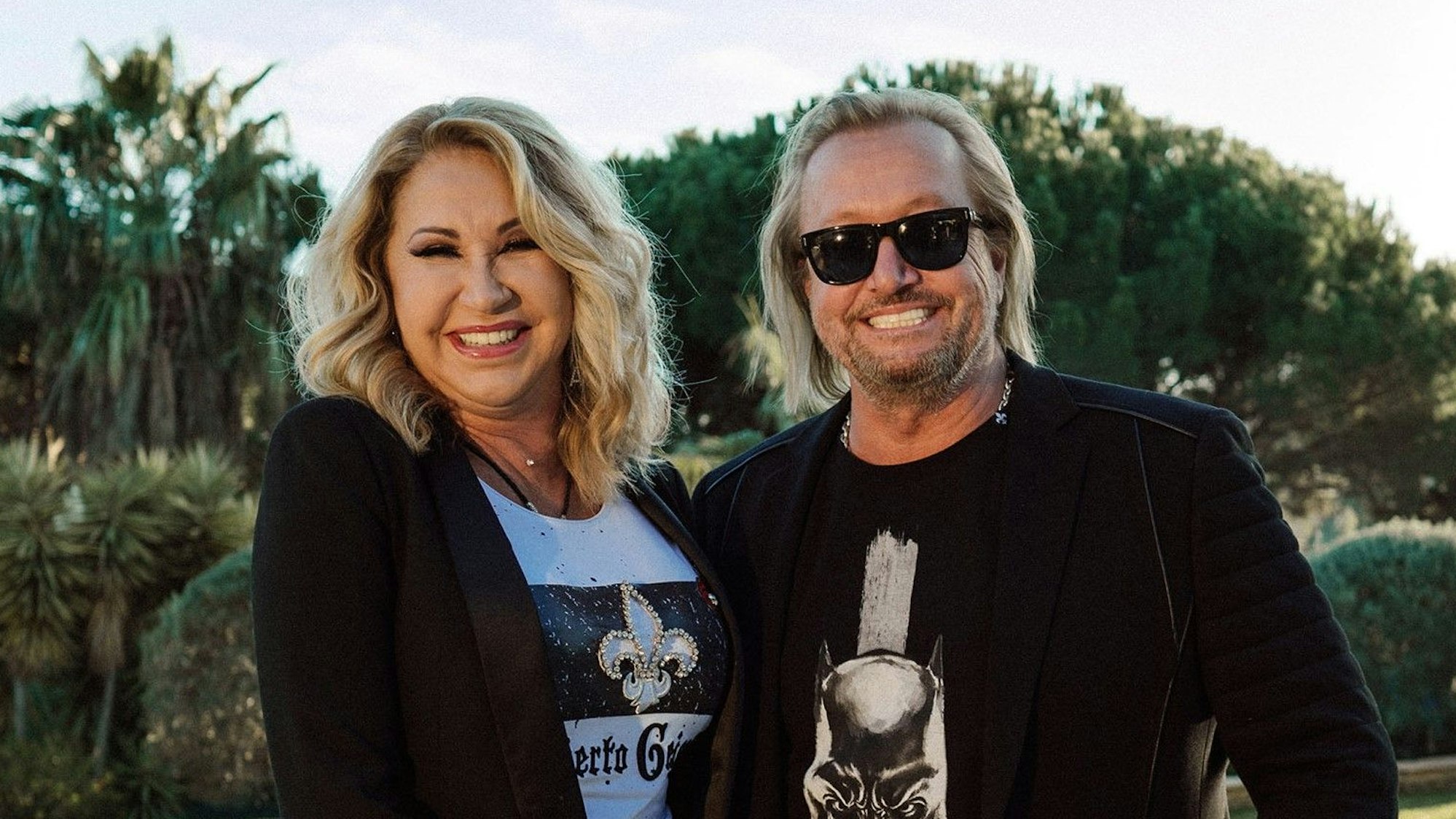 Carmen und Robert Geiss lachen posierend in die Kamera. Im Hintergrund ist die Natur zu sehen.